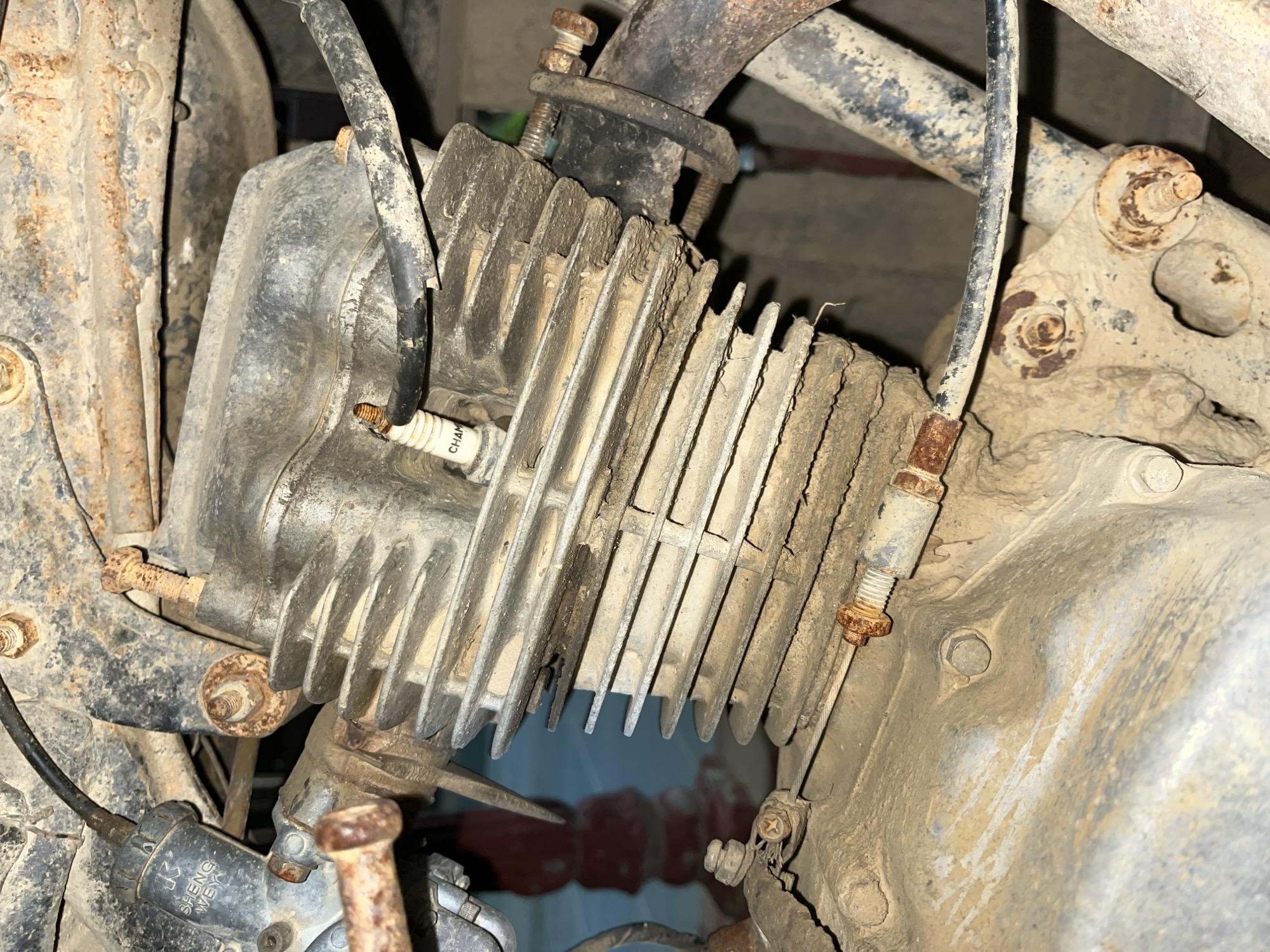 The image shows a close-up view of a dirty, possibly old, motorcycle engine. The main subject is the engine block, which is made of metal and has cooling fins. A spark plug is visible, screwed into the engine. There are various wires and cables connected to the engine, as well as some rusty bolts and brackets. The overall condition of the engine suggests it has been used extensively and is not well-maintained, with dust and dirt accumulated on its surfaces.
The setting appears to be a workshop or an outdoor area where the motorcycle is located, given the close-up nature of the shot focusing on the engine components. There is no indication of a specific time of day or weather conditions, and no people are present. The location context provided is Kota Kuala Simpang, Indonesia.
Notable details include the rust on several metal parts, particularly the bolts and brackets, indicating exposure to moisture or age. The spark plug is labeled with "CHAMPION". There is also some text visible on a black electrical component near the bottom left of the image, which appears to read "SHENG WEIYI". The image is taken from a slightly elevated angle, looking down at the engine. The focus is sharp on the engine components, while the background is blurred.