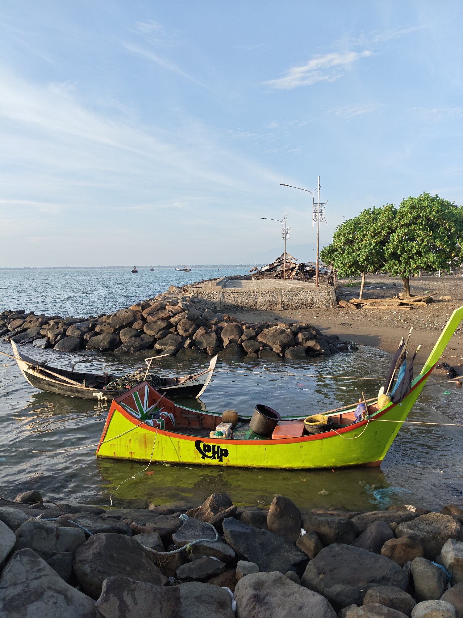 The image shows a coastal scene near Lhokseumawe, Indonesia. Two boats are the central focus. One boat is painted bright yellow with the letters "PHP" on its side, containing various items like buckets and nets. The second boat is smaller and darker in color. Both are partially submerged in the water near a rocky shore. The background features a small beach with a damaged structure, possibly a collapsed building, near a couple of light poles and a green tree. In the distance, there is a visible horizon with more boats on the water under a partially cloudy sky. The foreground features large rocks bordering the water. There are no people visible in the frame.