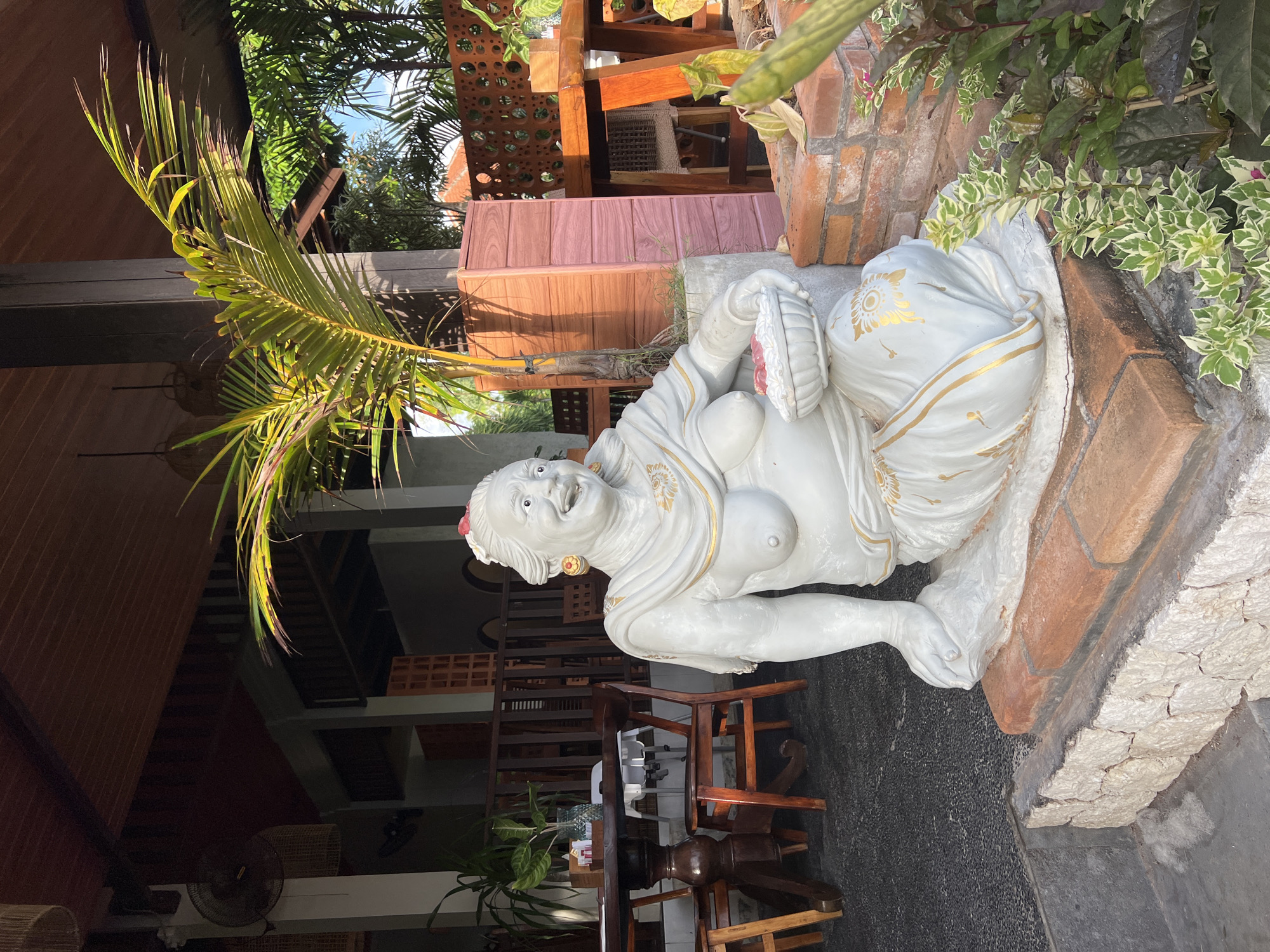 This image captures a vibrant, semi-outdoor scene, likely within a restaurant, cafe, or garden setting in Kuta Selatan, Indonesia. The central subject is a white statue of a reclining, corpulent woman, adorned with golden patterns on her drapery. She possesses a cheerful and jovial expression with an open mouth, and she holds a basket-like object in her left hand. The statue rests on a stone or concrete base, bordered by rustic red brickwork and surrounded by various plants, including a large palm frond in the foreground and background, as well as variegated green and white foliage.
The setting features tropical architecture with prominent wooden elements, such as beams, slatted screens, and reddish-brown paneling. Dark wooden tables and chairs are visible, arranged in what appears to be a dining or lounge area. A fan can be seen attached to a ceiling structure in the background, suggesting an open-air or semi-enclosed space. The ground is a dark, possibly tiled or paved, surface.
The image is taken during bright daylight, evidenced by the strong highlights and shadows, particularly on the palm frond and the statue, indicating sunny weather. The overall atmosphere suggests a relaxed, tropical environment. There is no visible text in the image.