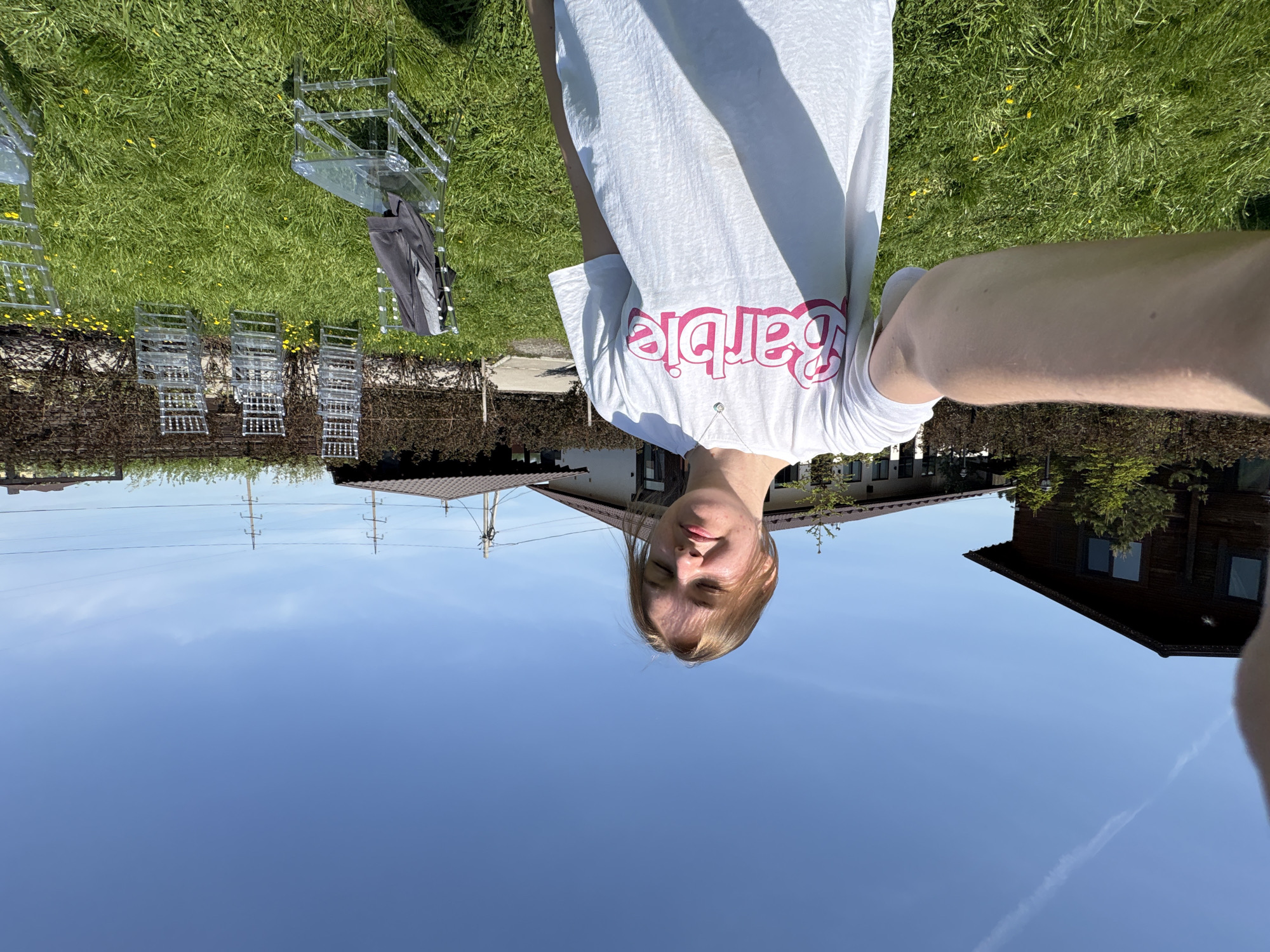 A topsy-turvy outdoor selfie captures a person wearing a white t-shirt with "Barbie" printed in pink, seemingly taken in a grassy area near a residential zone. The location appears to be Нижние Муллы, Russia, based on architectural and environmental cues present in the background. The sky is a clear blue, and several transparent chairs are positioned on the grass. Houses with gabled roofs are visible. Power lines are present in the background. The perspective is upside down.