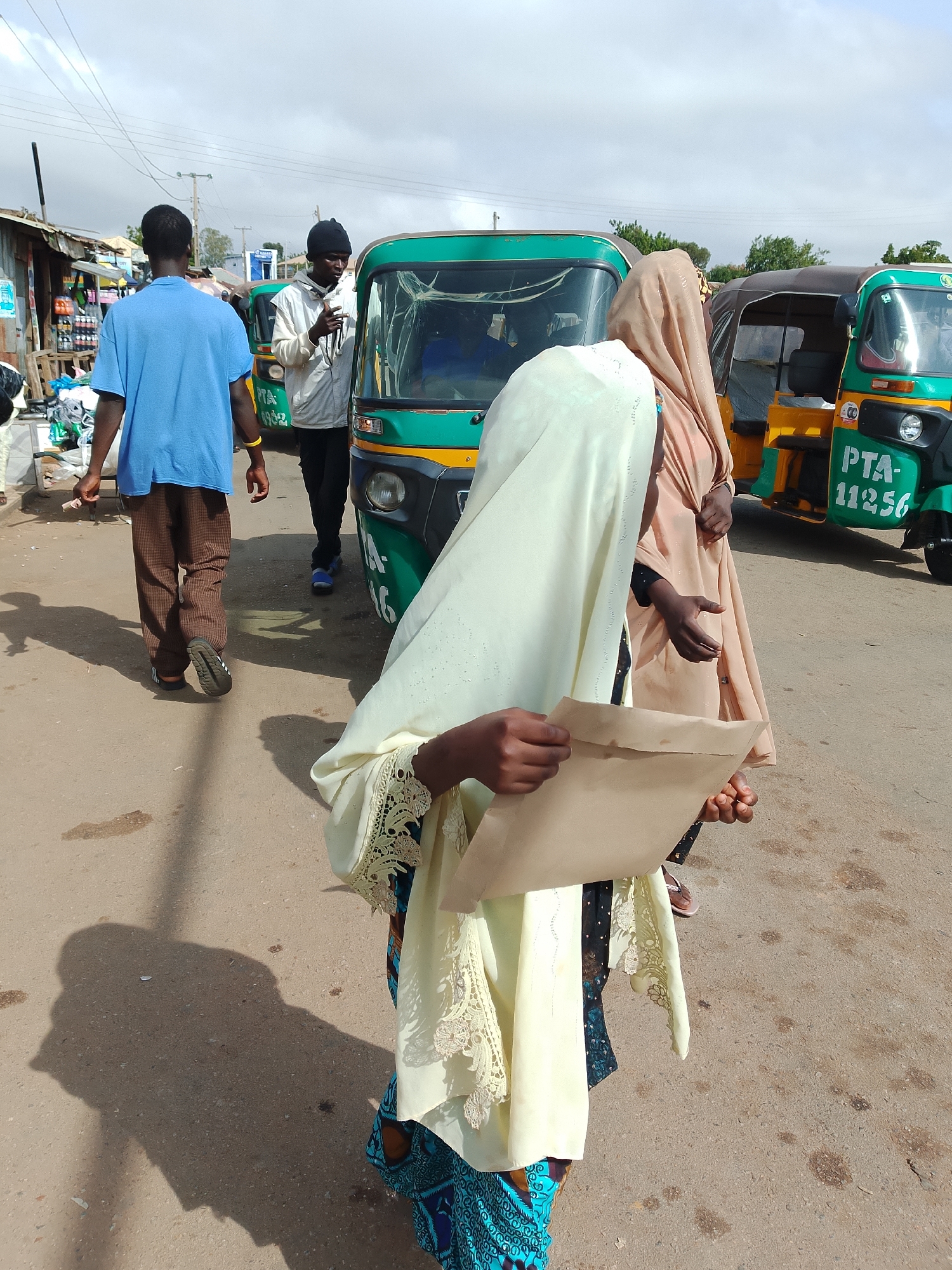 In Jos, Nigeria, a scene unfolds on a street with a weathered asphalt surface, hinting at a marketplace or transport hub. Power lines crisscross the hazy sky, above a row of informal stalls with corrugated iron roofing. Three-wheeled auto-rickshaws, locally known as "keke," are parked or moving along the street. One green keke, with the license plate "PTA-11256," is prominent. A young man in a blue t-shirt and checkered brown trousers walks away from the camera. Another man, wearing a black beanie and a white jacket, stands near a keke, holding a small object to his ear, possibly a phone. In the foreground, a person, likely a woman, is partially visible, draped in a pale yellow hijab and a patterned blue skirt, holding a brown paper envelope. Another figure, wearing a beige hijab, stands beside her. The overall atmosphere suggests a bustling, everyday moment in this Nigerian city.