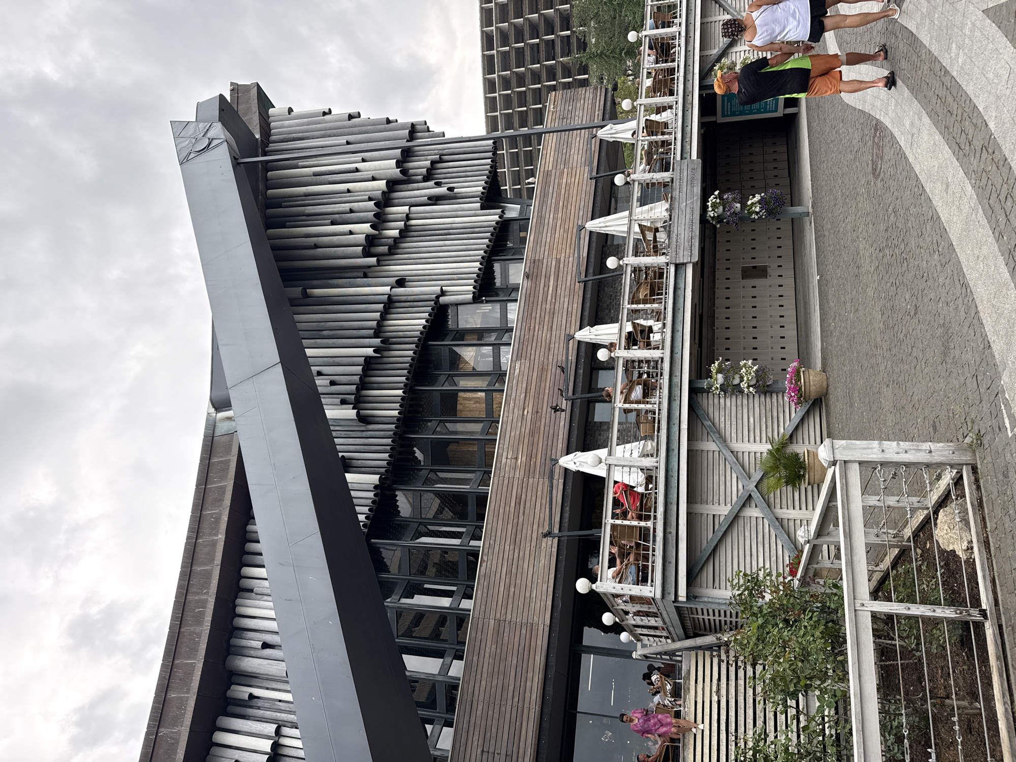 This is a medium-shot, eye-level outdoor photograph of the exterior of a building with a prominent, angular grey structure extending from its roofline. The building itself is multi-layered, with a wooden-paneled section on the second level featuring a balcony or terrace with tables, chairs, and white umbrellas. Several people are seated at these tables. A woman in a pink dress is walking down a set of stairs on the left side of the building, while two people, a man in an orange hat and shorts and a woman in a white tank top and black shorts, are walking away from the camera on a paved pathway to the right. The background shows a dense grid-like structure, possibly another building or part of the same complex, and some greenery. The sky is overcast. There is a sign on the building that appears to be in Cyrillic and English, with words like "Storage." The scene is set in the city of Yalta, Ukraine.
