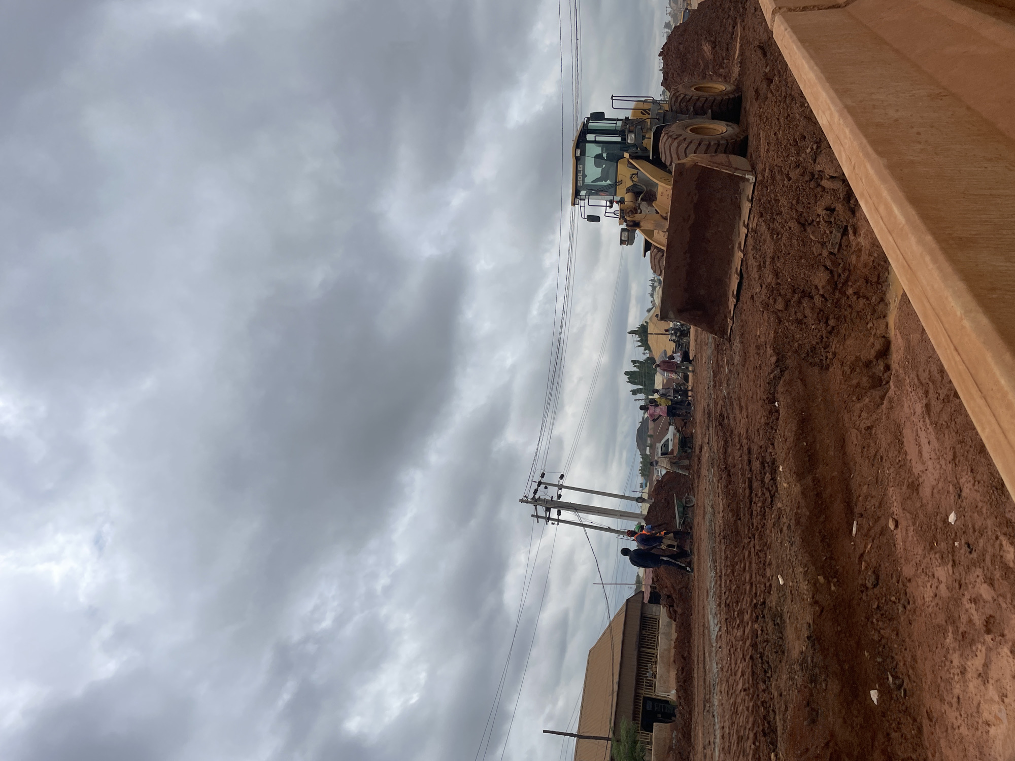 A yellow front-end loader is parked on a construction site, its bucket filled with reddish-brown dirt. The scene is in Minna, Nigeria. The sky is overcast with dark, dramatic clouds. In the background, several people are working, some with wheelbarrows, and there are buildings and utility poles visible. The ground is uneven and covered in dirt, indicating ongoing construction or roadwork. A temporary concrete barrier is in the foreground.