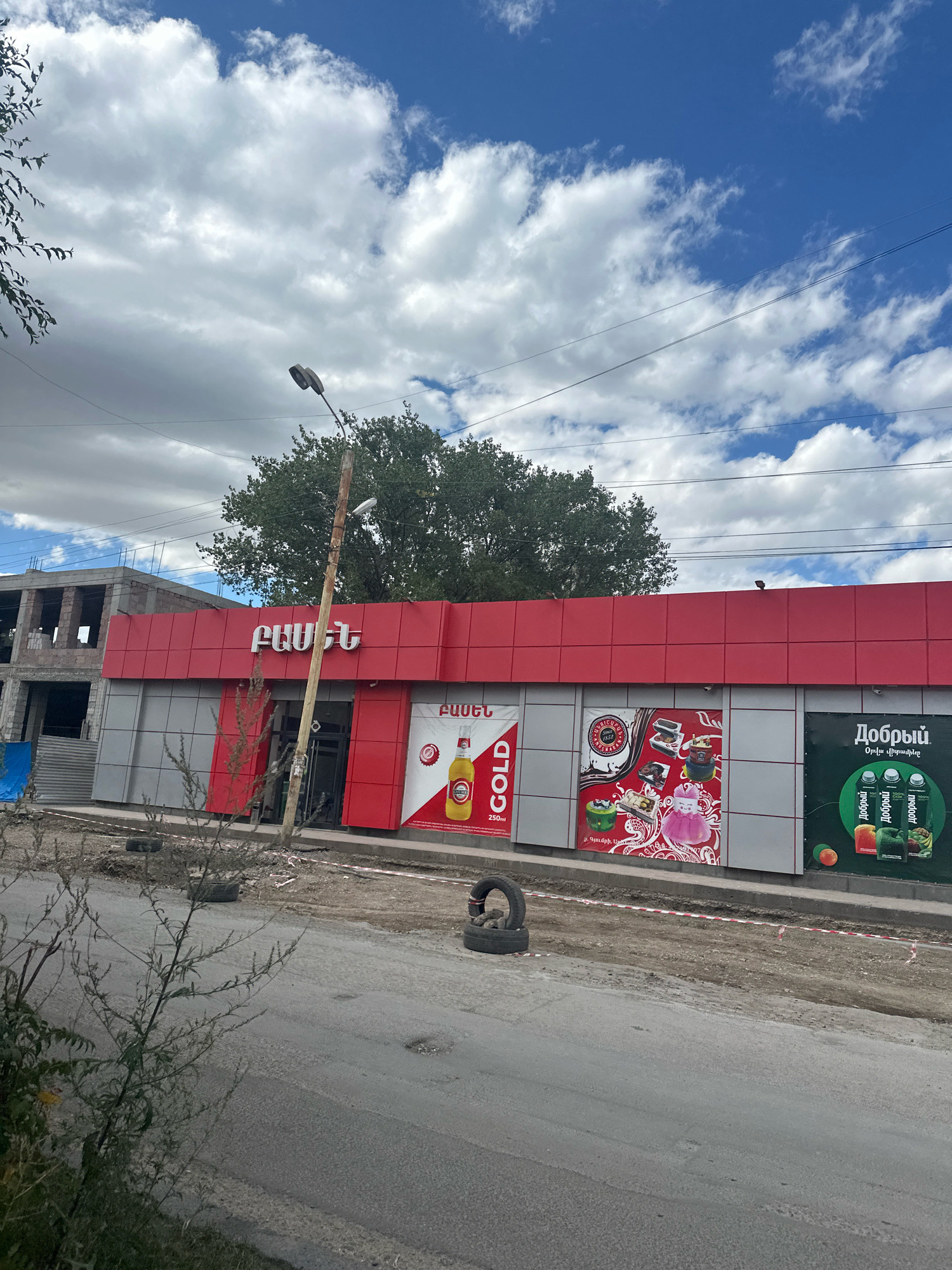 The image shows a street scene in Gyumri, Armenia. In the foreground, there is a road with some construction materials and debris. A small, leafless bush is growing on the left side of the road. In the midground, there is a modern-looking building with a red and silver facade. The building appears to be a shop or store, with large advertisements displayed on its exterior. One advertisement features a bottle of "GOLD" drink, another showcases various food items, and a third displays bottles of "Добрый" juice. The building is also under construction on the left side, with visible concrete structures and rebar. A lamppost stands in front of the building. In the background, there are trees and a cloudy sky with blue patches. The overall scene depicts an urban environment undergoing development or renovation.