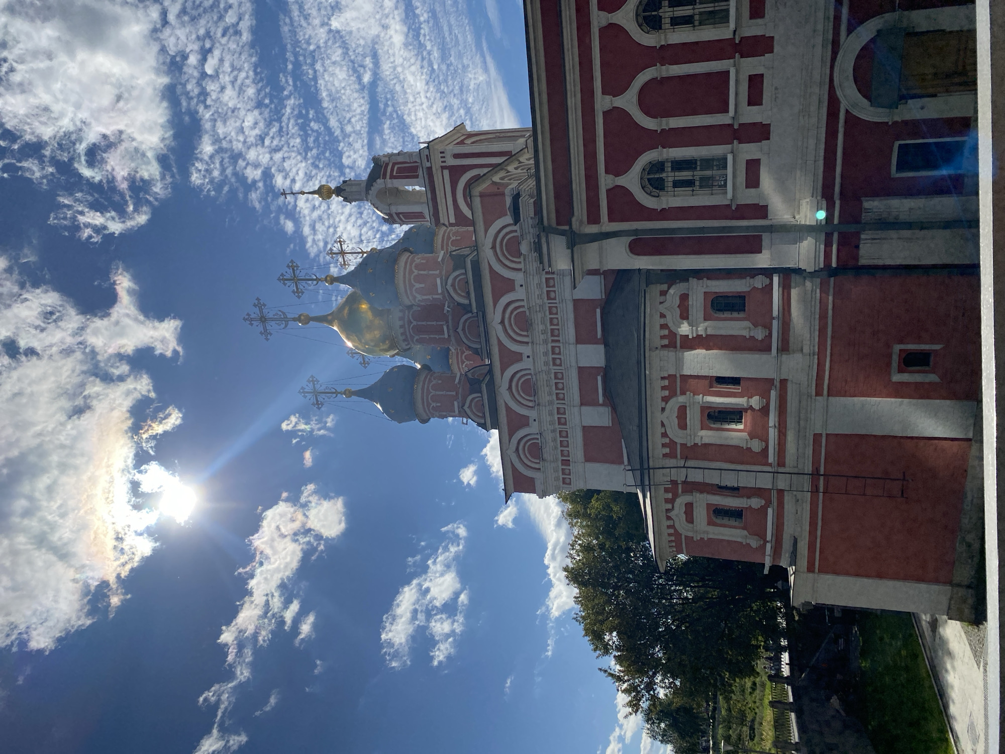 The image shows a view of the Church of the Ascension of the Holy Virgin in Kolomenskoye, Moscow, Russia. The church, with its distinctive red and white facade and multiple blue and golden domes topped with crosses, is bathed in sunlight. The sky is a vibrant blue with scattered white clouds, some of which are iridescent due to the sun's glare. In the foreground, a portion of a grey railing is visible, suggesting the photo was taken from an elevated vantage point. To the left of the church, lush green trees are visible, and beyond them, a stone bridge with people walking across it can be seen. The overall scene depicts a sunny day with a clear view of the historic church and its surroundings.