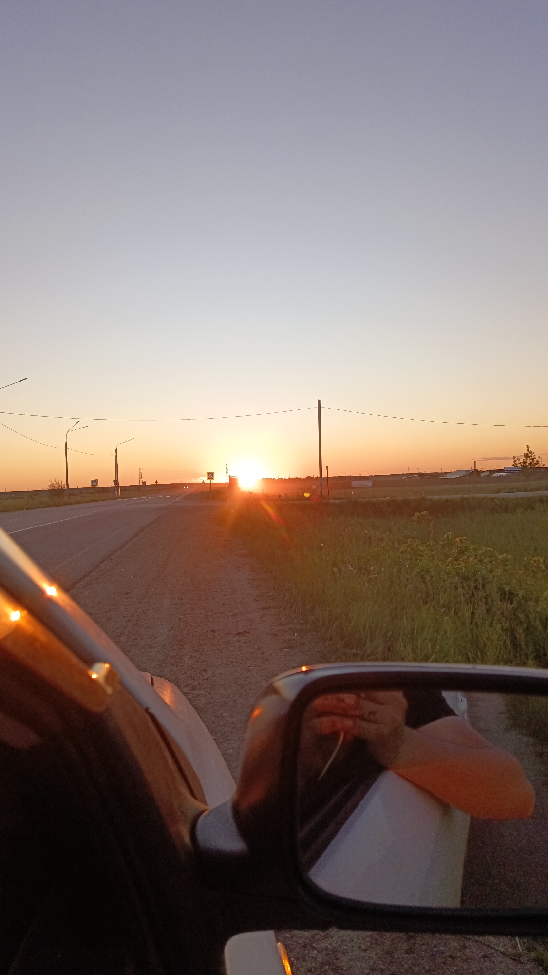 The image depicts a sunset scene viewed from the perspective of a moving vehicle on a rural road. The sun is low on the horizon, casting a warm, orange glow across the sky and landscape. The road stretches into the distance, lined with grassy fields and sparse vegetation. In the foreground, a side-view mirror reflects a person's arm and hand, possibly resting on the car's interior. The overall atmosphere is serene and captures the beauty of a sunset in a tranquil, natural setting.