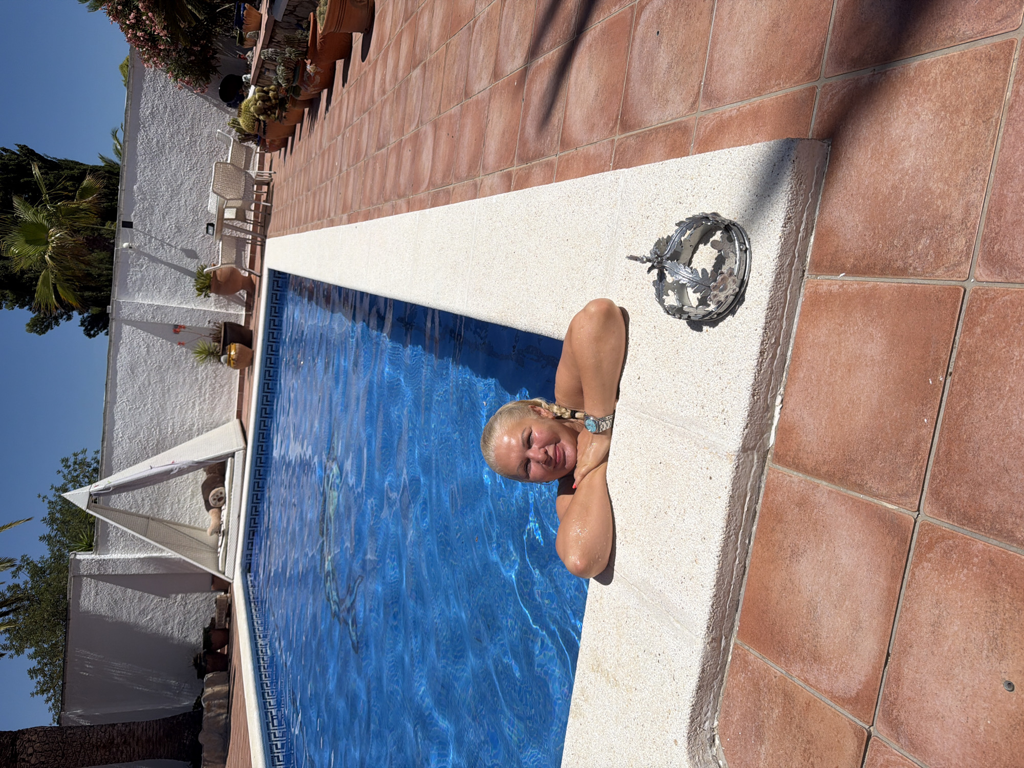 In Mont-roig del Camp, Spain, a woman with blonde hair pulled into braids is partially submerged in a bright blue swimming pool, leaning on the pool's edge. She is wearing a watch on her left wrist and a turquoise pendant on a chain. Next to her, on the pool's edge, is a small, ornate silver crown with leaf motifs. The pool is surrounded by a red tiled patio. In the background, a white stucco wall encloses the property, with a triangular white cabana containing cushions and a rolled-up mat. Various potted plants, including cacti, and patio furniture are visible along the wall. The sky is clear and blue, indicating a sunny day.