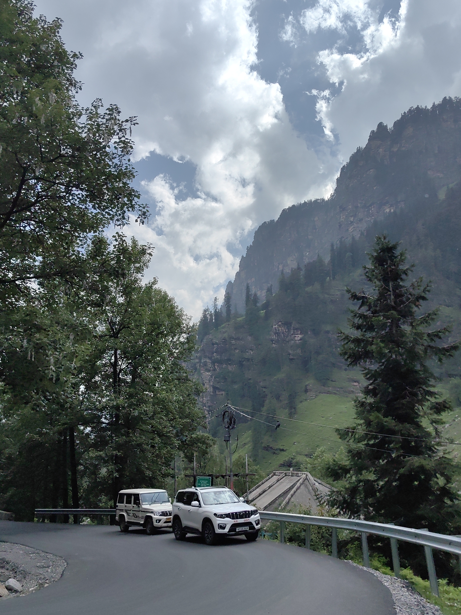 In Kothi, India, two white SUVs are on a paved road that curves to the right, bordered by a metal guardrail. The SUV in the foreground is white and has a license plate that reads "HP 33G 5808". Behind it is another white SUV, a Mahindra Bolero. Tall green trees flank the left side of the road, and a large, rocky mountain covered in trees dominates the background. The sky is filled with dramatic, white clouds.