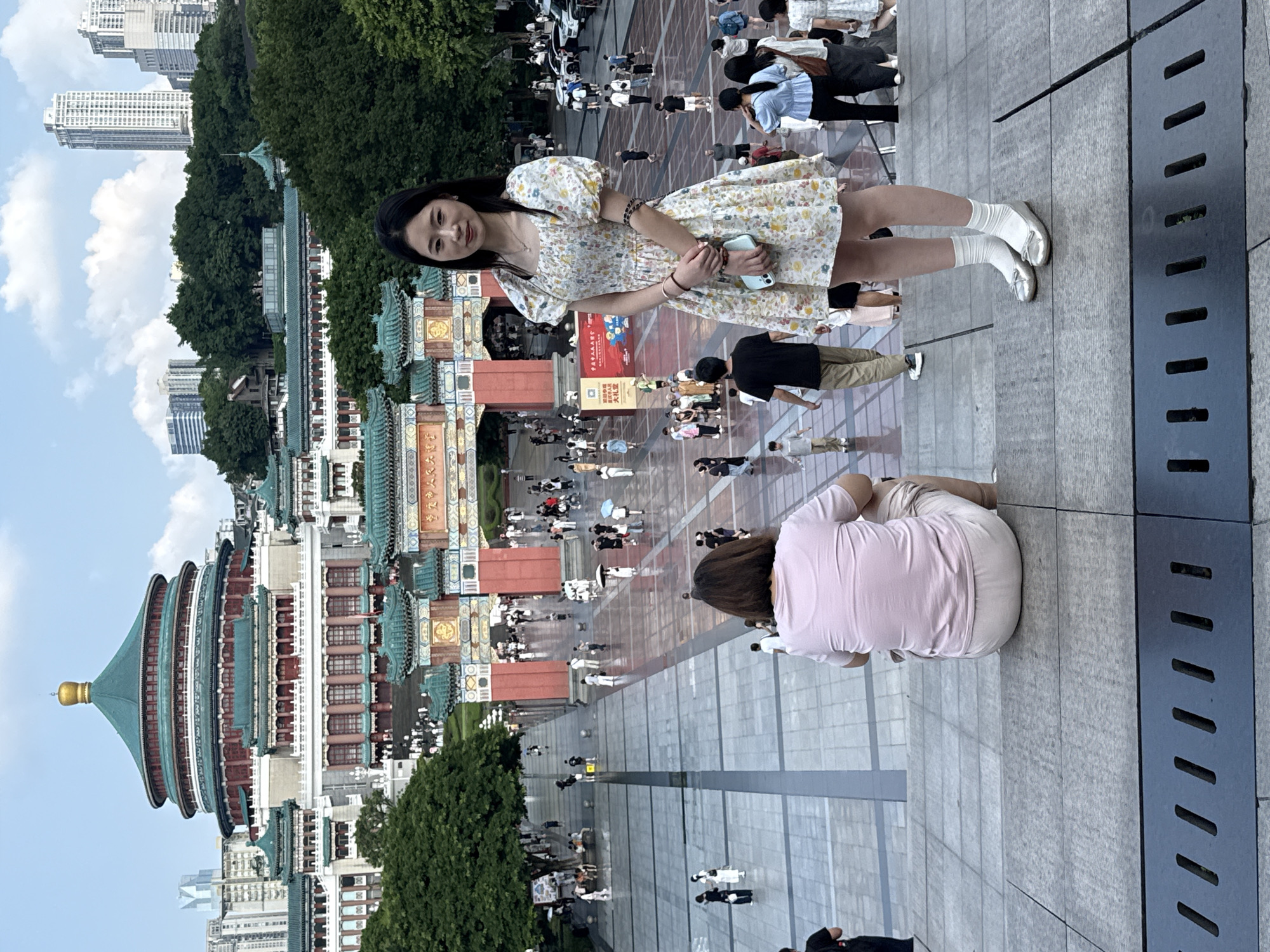 In Yuzhong District, China, a young woman in a floral dress stands on stone steps, holding a smartphone. Her silver ballet flats and white socks contrast with the grey steps. Behind her, a person sits facing away, wearing a pink t-shirt and light-colored shorts. In the background, a large, ornate building with a green, domed roof and red accents dominates the scene, flanked by traditional Chinese architecture with green-tiled roofs. Numerous people are dispersed across a wide, paved plaza leading up to the building, some walking, others standing or sitting. Trees and modern high-rise buildings are visible in the distance. The sky is partly cloudy.