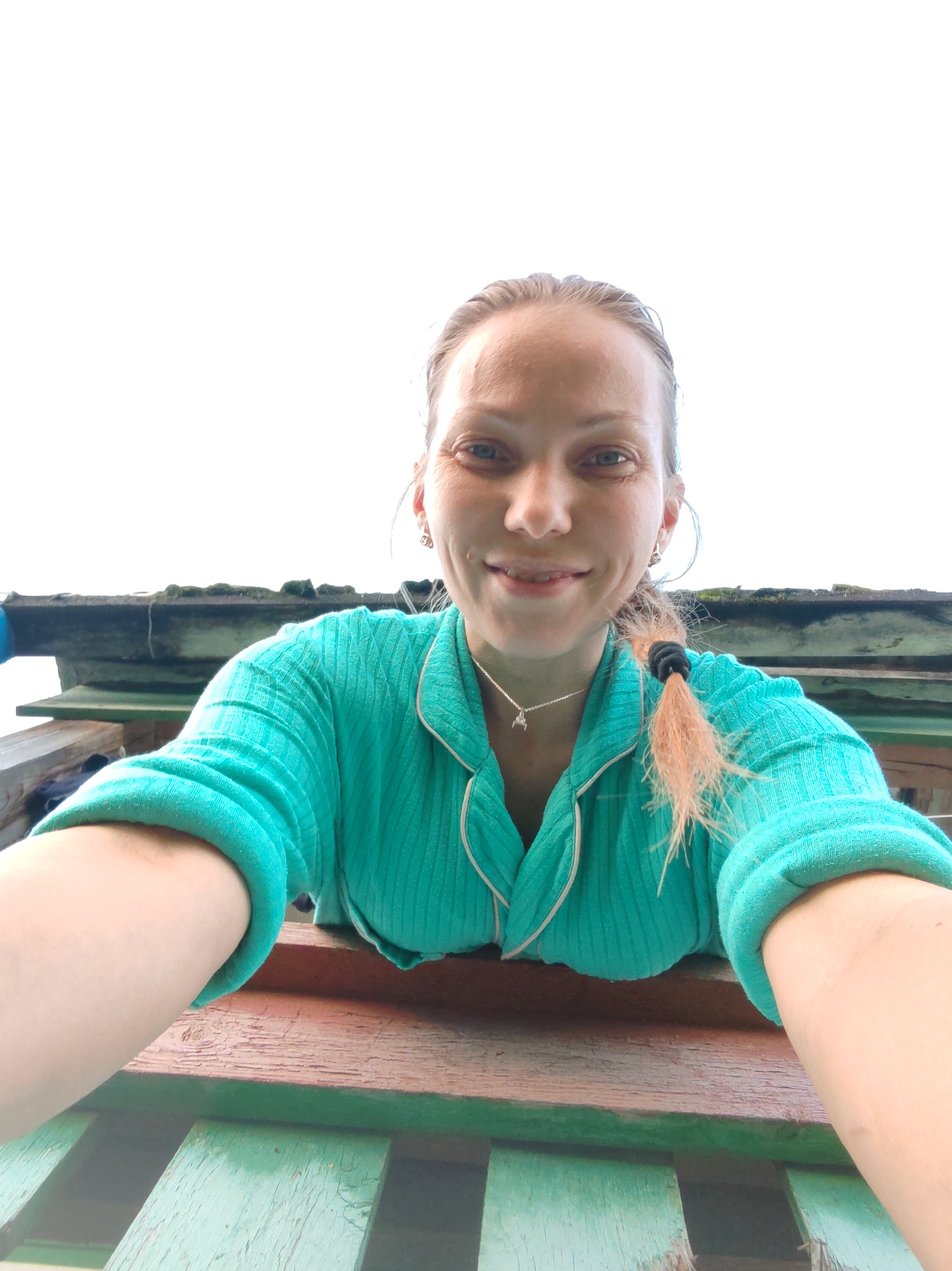 In the city of Chukhlem, Russia, a young Caucasian woman with blonde hair pulled back in a braid and wearing a teal pajama set is captured in a selfie. She is leaning over a wooden railing, with a weathered roofline and structure visible in the background. The sky is bright white, suggesting it is daytime. The woman is smiling and looking directly at the camera. The image is taken from a low angle, looking up at her.