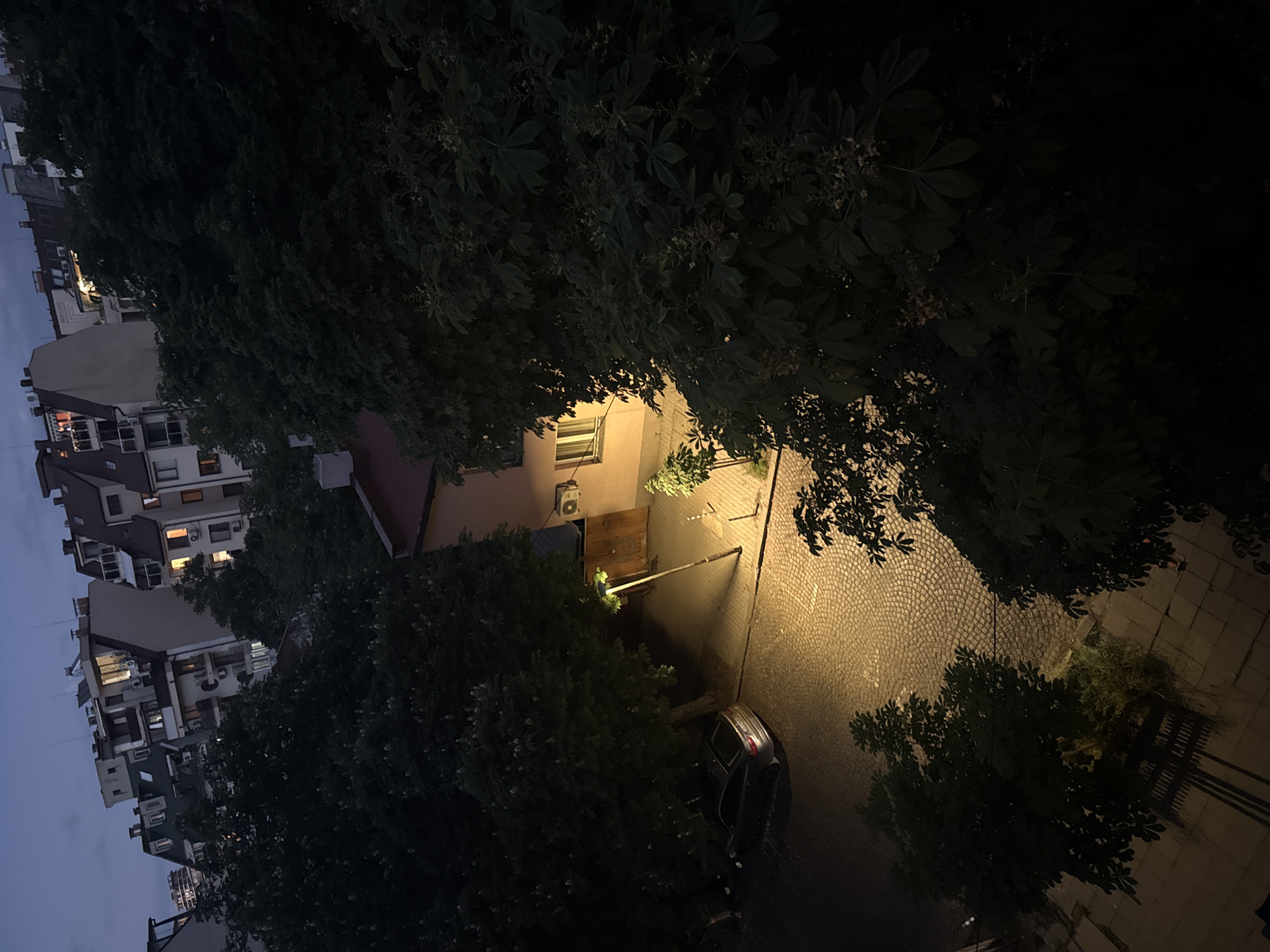 An aerial view, from an elevated position, shows a street scene in Sofia, Bulgaria at night. A narrow cobblestone street, illuminated by a street lamp, cuts diagonally through the frame, receding into the distance. Mature trees with dense foliage frame the street, partially obscuring the buildings and the night sky. Residential buildings with multiple floors and various architectural styles can be seen along the left side of the frame. A car is parked on the right side of the street. A building with a light brown facade and an air conditioning unit mounted on the exterior wall stands out amidst the surrounding buildings.