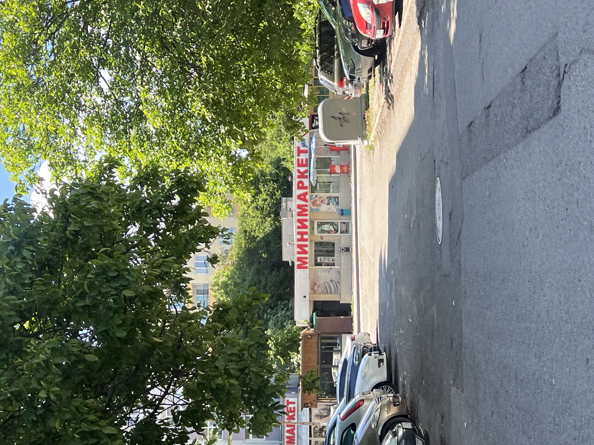 The image shows a street in Burgas, Bulgaria, featuring a small minimarket. The building has "МИНИМАРКЕТ" written vertically on the front. The minimarket is situated between parked cars and under the shade of trees. The street has a dark asphalt surface. The environment includes trees, other buildings in the background, and various parked cars, creating a typical urban street scene.