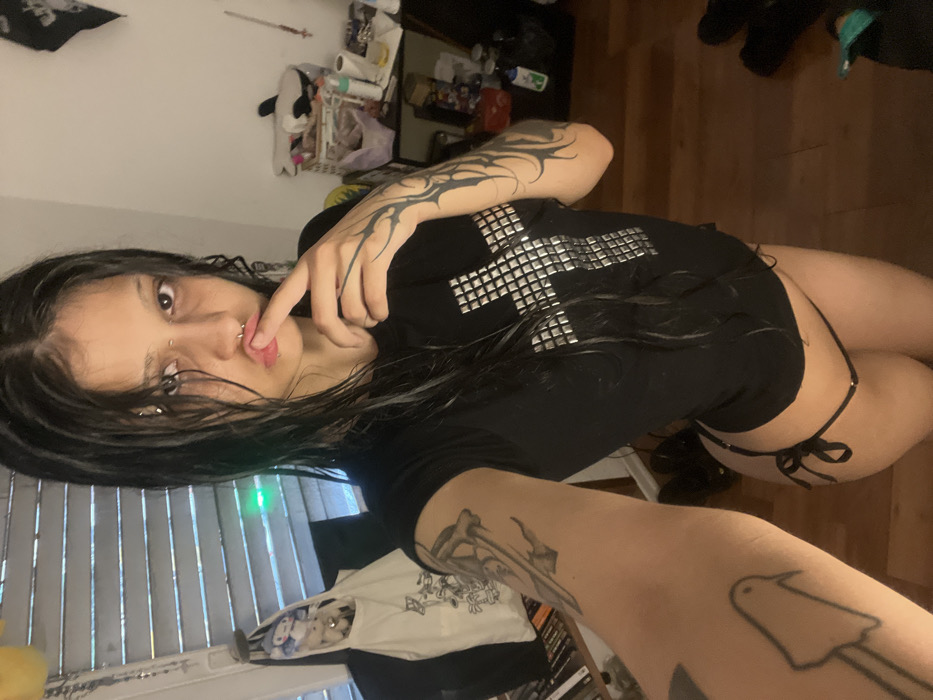 A young woman poses for a selfie in a cluttered room. She wears a black T-shirt with a studded cross design and a black strappy bottom. She has dark hair, pierced lips and nose, and extensive tattoos on her arms. Blinds cover the window behind her, and items are piled on a shelf in the background. The floor is wooden.