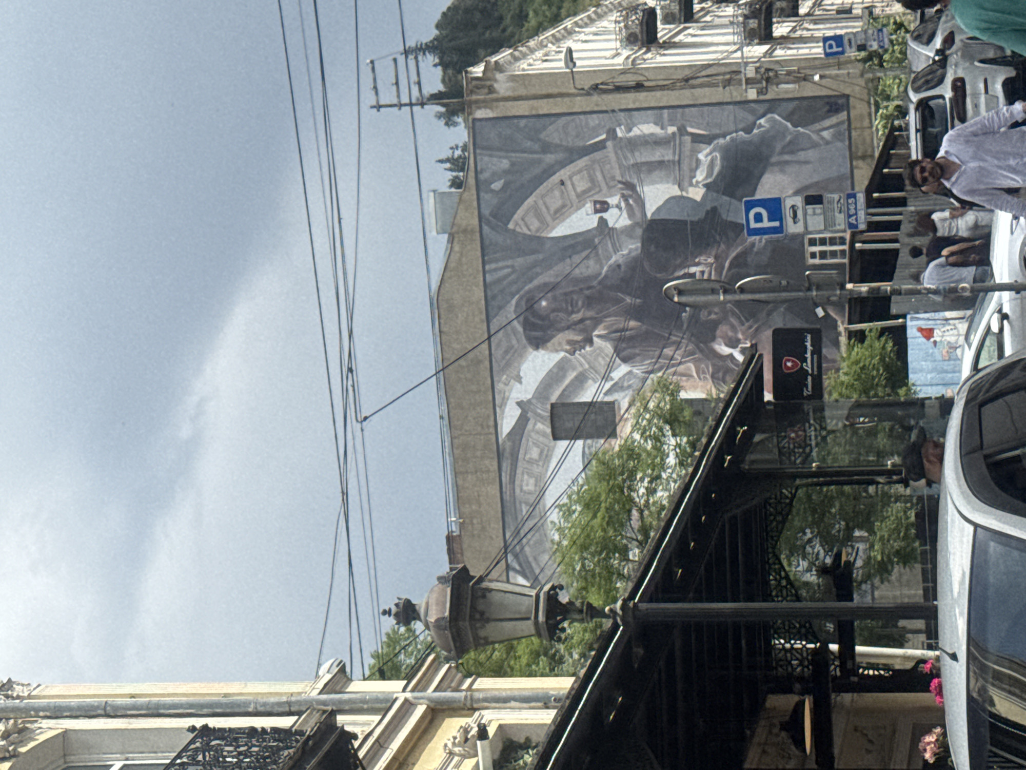 This is a medium shot of a street in Tbilisi, Georgia, taken from a low angle looking up. The sky is overcast with dark clouds. The main subject is a large mural on the side of a building, depicting two figures in profile, one holding a glass. The mural is rendered in shades of gray and appears to be set in an arched, architectural background.
In the foreground, a black canopy structure extends from the building on the left. A lamppost with ornate detailing stands in front of the canopy. To the right of the mural, a multi-story building with white facade and balconies is visible. A street light is mounted on this building.
On the street level, several cars are parked. A white car is in the lower left foreground. Further down the street, other white cars and a man in a light-colored shirt walking towards the camera are visible. A blue parking sign with a "P" is also present.
Trees are visible behind the canopy and on the hillside in the background, suggesting a vegetated urban environment. The overall impression is of a slightly gloomy, cloudy day on a city street adorned with street art.