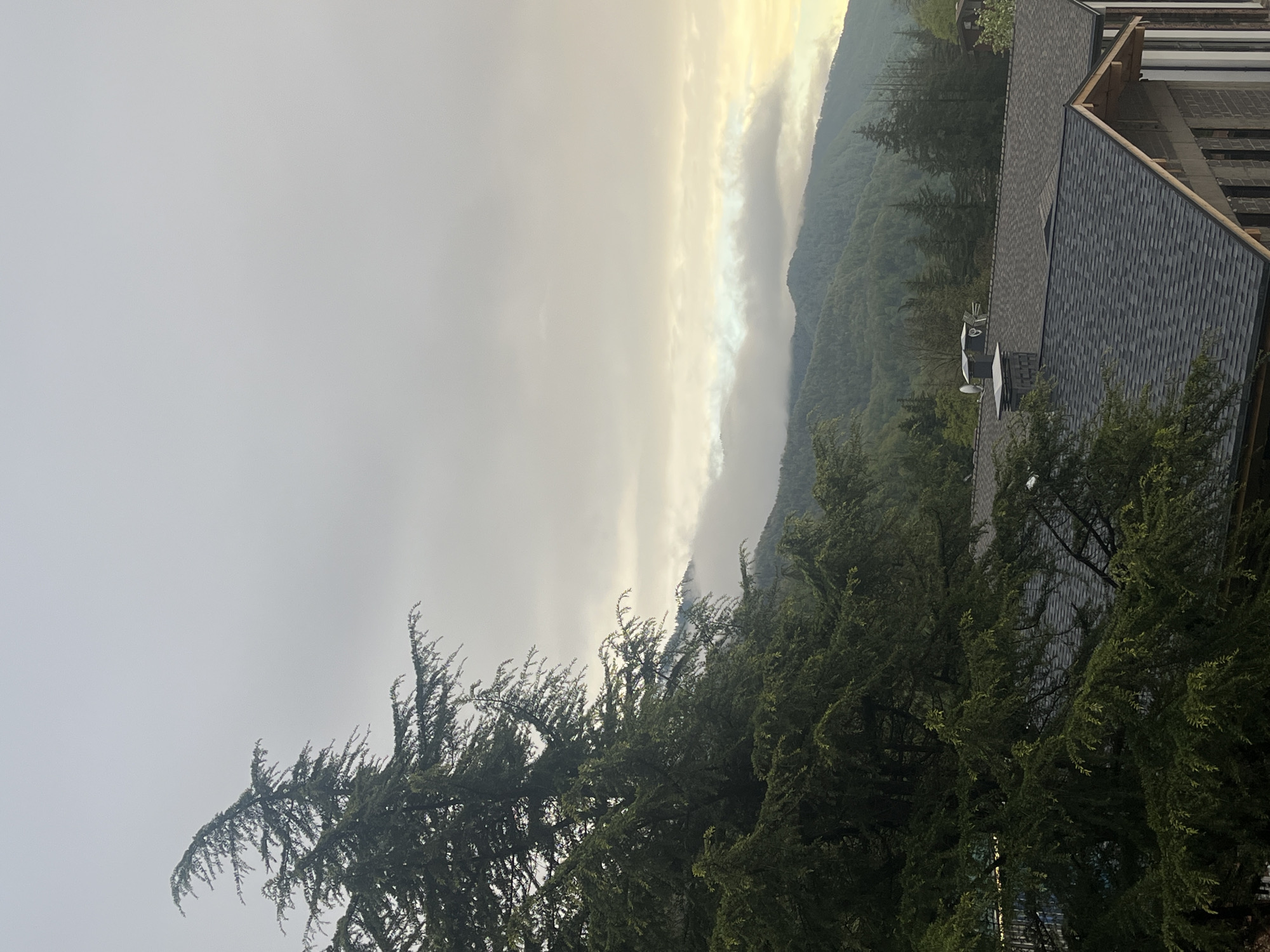 The image depicts a landscape with natural and built elements, possibly in a mountainous or hilly region. The sky is overcast with varying shades of gray and some lighter areas suggesting a break in the clouds or the sun filtering through. Mountainous terrain is visible in the background, partially obscured by clouds or fog. The middle ground features dense green trees, likely evergreen species. A portion of a building with a gray shingled roof and metal fixtures is present in the upper right corner. The location is unknown.
