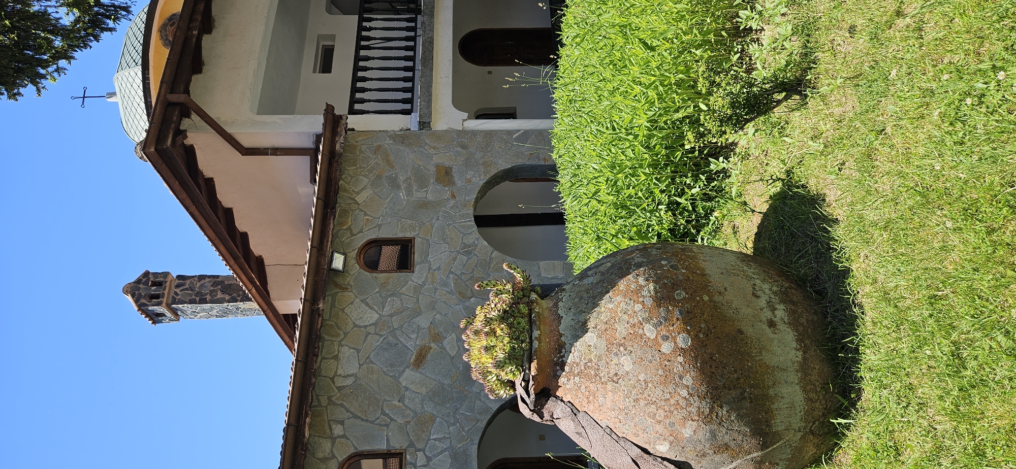 The image shows an outdoor scene with a large, weathered clay pot in the foreground planted with succulents. Behind the pot is a building constructed with stone walls and a section with white plaster. The building has arched doorways and windows, a balcony with a dark railing, and a domed roof topped with a cross. A stone chimney with multiple openings is visible on the roof. Lush green foliage surrounds the building and extends to the right of the pot. The sky is clear and blue. The scene is set in the village of Ovchartsi, Bulgaria, likely in the vicinity of the Ovchartsi Monastery, a notable landmark in the region. There are no people or animals present in the image.