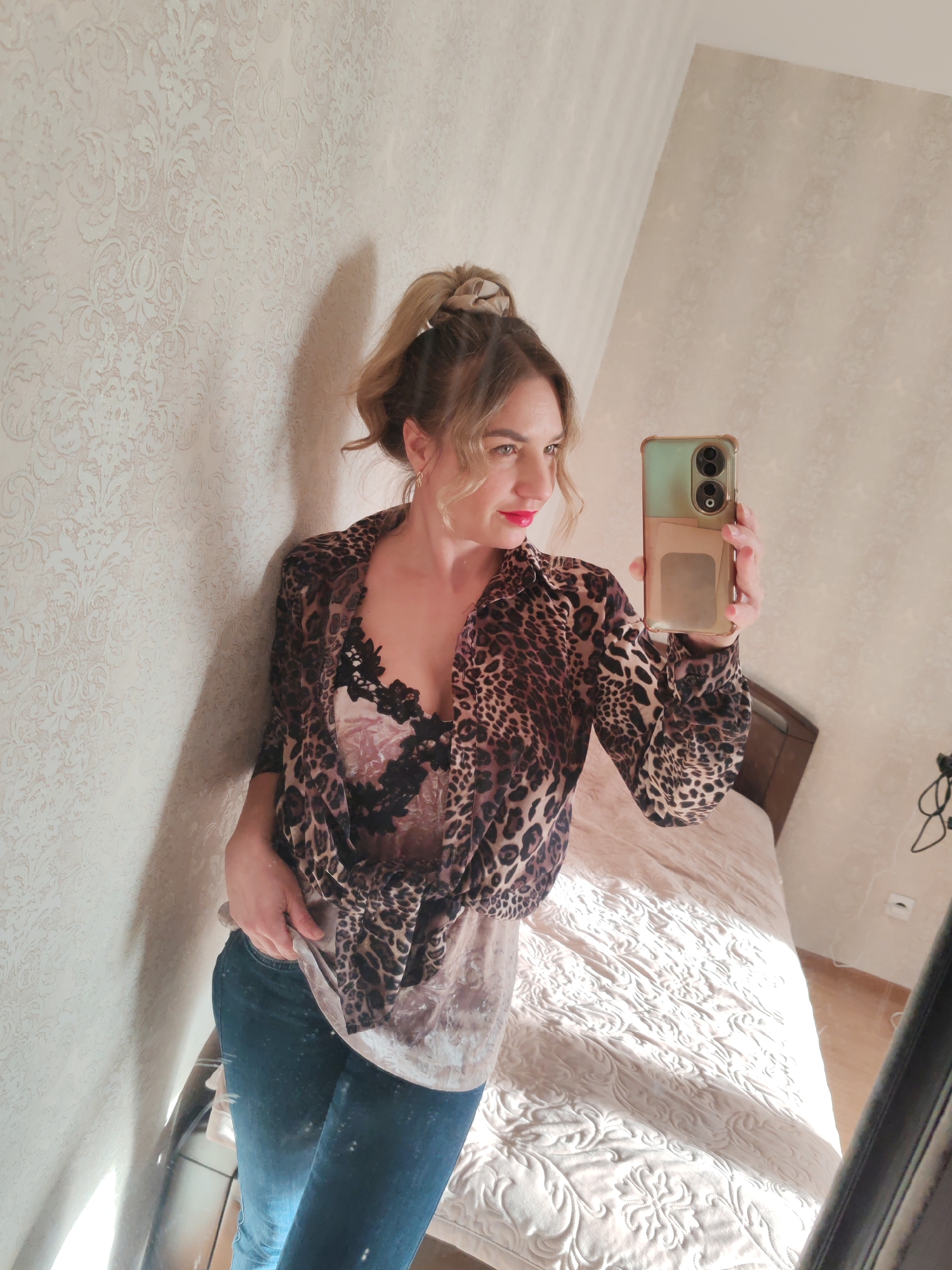 A woman is taking a mirror selfie in a room with light beige wallpaper. She is wearing a leopard-print shirt over a black lace top and blue jeans. Her hair is pulled back in a high ponytail with a beige scrunchie. She is holding a smartphone with a two-toned case. A bed with a light-colored patterned comforter is visible in the background. The location is not specified.