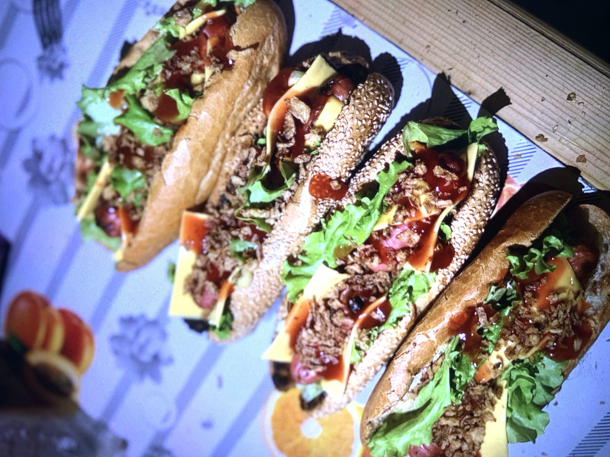 The image presents a close-up, overhead view of five elaborately prepared hot dogs or sausage sandwiches arranged diagonally on a light blue and white striped placemat. To the right, a wooden surface, likely a table, is visible.
The hot dogs showcase a variety of buns; some are traditional light brown rolls, while others are darker and generously coated with sesame seeds. Each sandwich is filled with a sausage, bright yellow cheese slices, fresh green lettuce, and a topping resembling crispy fried onions or seasoned crumbled meat. A rich red sauce, possibly ketchup or a similar condiment, is generously drizzled over the ingredients.
The lighting appears artificial, casting noticeable deep shadows beneath the hot dogs and the wooden edge, suggesting an indoor setting. In the blurred foreground on the left, two stacked, round, orange-red objects, possibly fruit slices or decorative items, are faintly discernible, along with a blurry pattern on the placemat that hints at a citrus slice design.
The scene captures food prepared for consumption, indicating a casual meal or snack. There are no individuals present, nor is any visible text. While the location is contextualized as Severodvinsk, Russia, no specific visual elements within the image confirm this geographical detail.