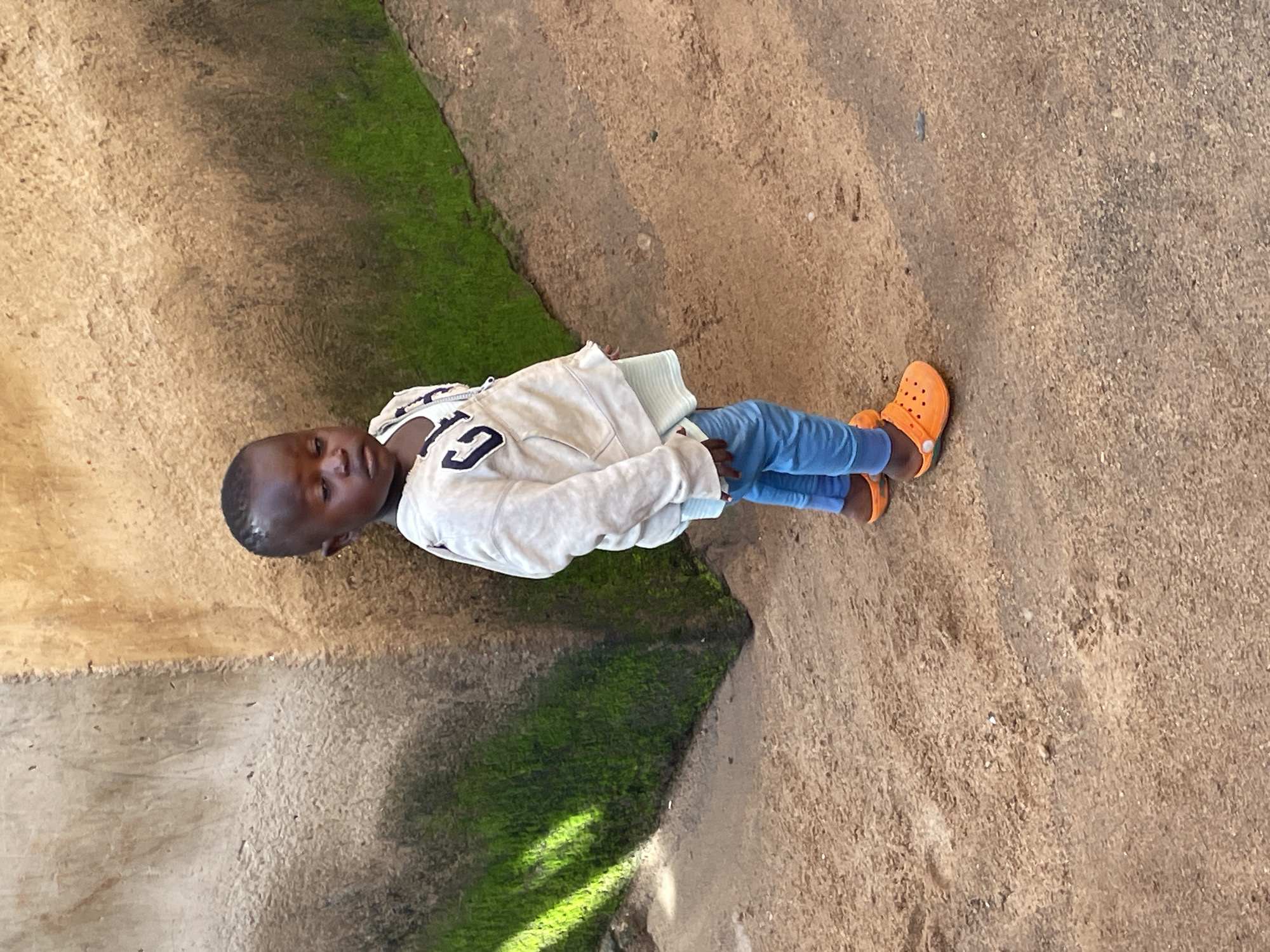 A young child stands on a dirt path in front of a textured, beige wall with patches of green moss. The child is wearing a light-colored hooded sweatshirt with "GAP" logo in navy blue, light blue pants, and bright orange Croc-style shoes. The child is looking downwards, with their head tilted slightly. The wall appears to be part of an outdoor structure. The lighting suggests it is daytime, with shadows cast on the ground. The overall scene is likely in an outdoor, possibly rural or less developed area.