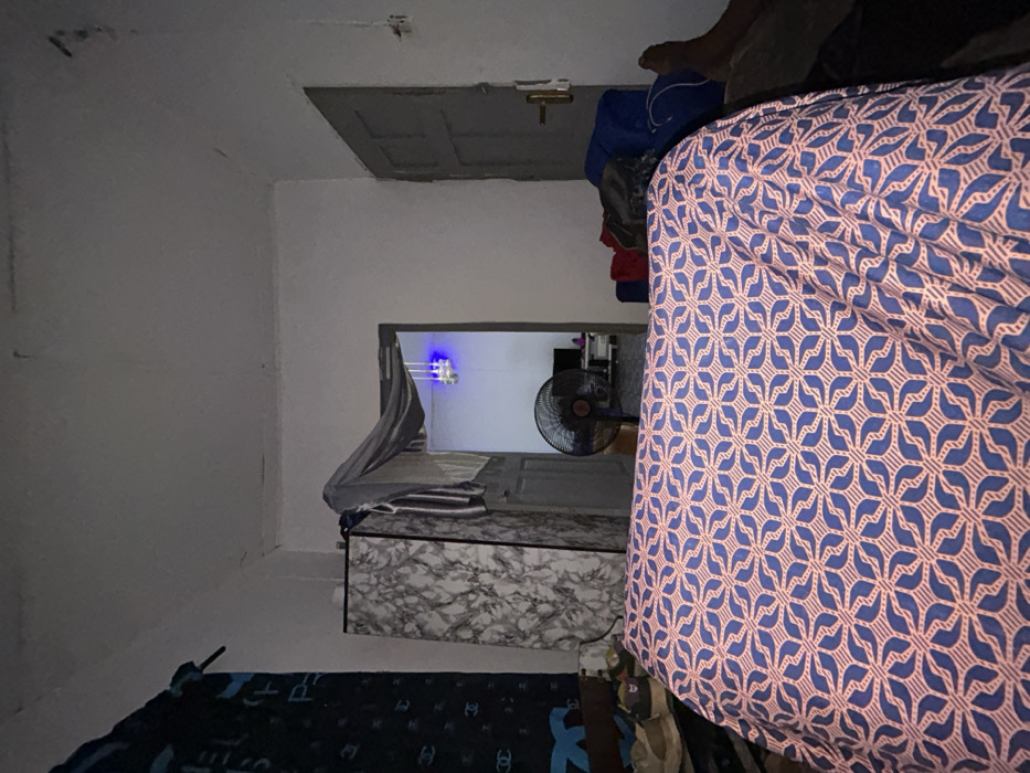 The image depicts the interior of a dimly lit room in Ilishan, Nigeria, viewed from a low perspective, likely from a bed. The foreground is dominated by a bed covered with a light pink/orange sheet featuring a repetitive blue geometric pattern of intersecting lines and stylized leaf motifs.
To the left, a dark blue curtain with light "CHANEL" logos hangs partially. Below it, several pairs of shoes, including light brown sneakers, rest on a surface. Adjacent to this, a tall, rectangular cabinet or wardrobe with a grey and white marble-like pattern stands. A light grey fabric and other clothes, including a striped item, are draped over the cabinet's top edge.
In the center, a doorway opens into an adjacent room. A black standing fan is positioned in this doorway, its circular cage visible. Inside the further room, a bluish light source hangs from the ceiling, and a dark rectangular object, possibly a television, is visible on a surface in the background. The floor in the second room appears to be tiled.
To the right of the main room, a closed grey door with a light-colored handle is present. Near the bottom right corner, a dark blue bag, possibly a duffel bag, is placed on the floor alongside some red fabric. A bare human foot, likely the right foot, is visible in the extreme lower right, resting on a dark surface next to the bed. The walls are light-colored, showing some discoloration and minor imperfections, particularly near the ceiling, which also exhibits some marks. No direct human interaction is depicted, but the presence of the foot suggests an individual is within the immediate vicinity.