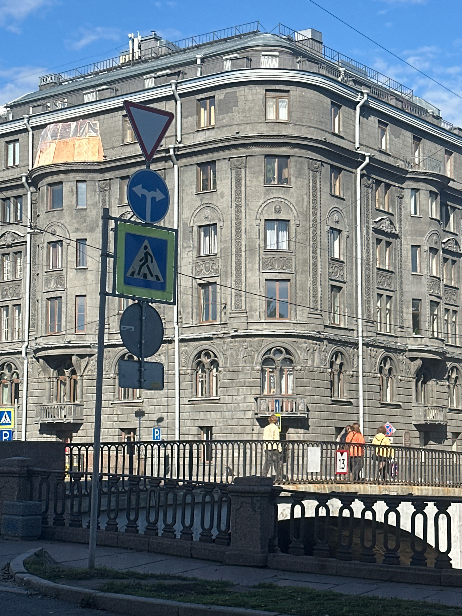 The image shows a view of the Anichkov Bridge over the Fontanka River in Saint Petersburg, Russia. The iconic bridge is identifiable by its distinctive railings and the historical building facade visible in the background. Several traffic and pedestrian signs are present, including a yield sign, a pedestrian crossing sign, and a blue circular sign indicating direction. On the bridge, three individuals are walking, two of whom are wearing brightly colored jackets. The architecture of the building in the background is classical with ornate details and multiple windows. The sky is blue with some clouds, suggesting a pleasant day. The foreground shows a sidewalk and a grassy area.