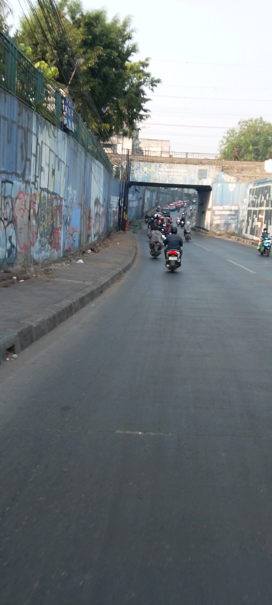 The image depicts a street scene in Bekasi, Indonesia, characterized by a road with moderate traffic, primarily consisting of motorcycles. A concrete wall with graffiti lines the left side of the road, separating it from a pedestrian walkway. Beyond the wall, trees and vegetation are visible. An overpass or bridge spans the road in the middle ground, with traffic visible on it as well. The overall atmosphere suggests a typical urban environment in Indonesia, likely during daylight hours due to the clear sky and visible shadows.