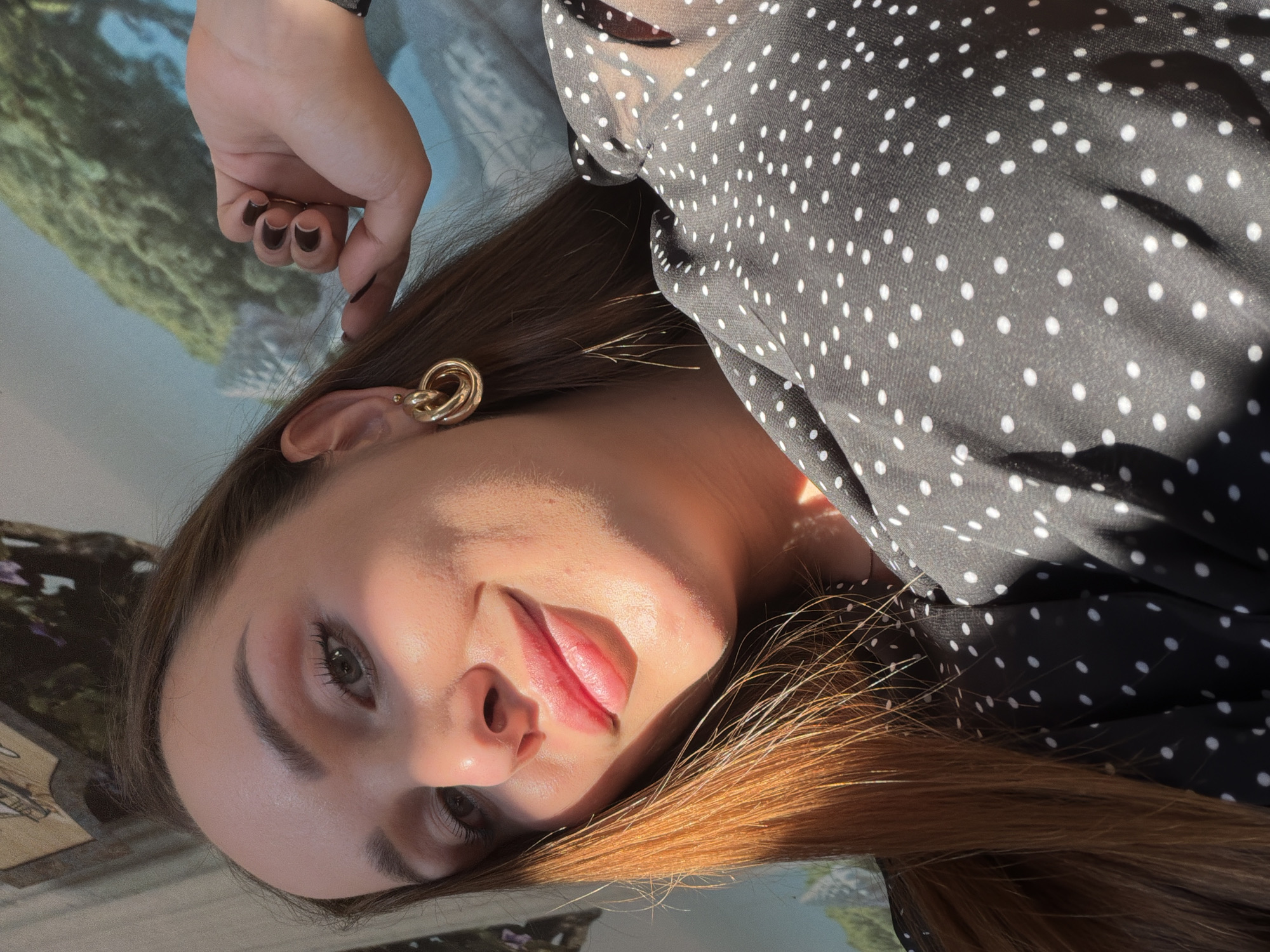 A medium shot captures a young woman with long, auburn hair indoors, bathed in sunlight. She is wearing a black dress with white polka dots and large, gold hoop earrings. Her right hand is resting near her face, with dark nail polish visible. The background is blurred, suggesting a wall with a mural or artwork depicting a landscape. The lighting creates strong shadows on her face, highlighting her features. The woman is looking towards the camera with a slight smile.