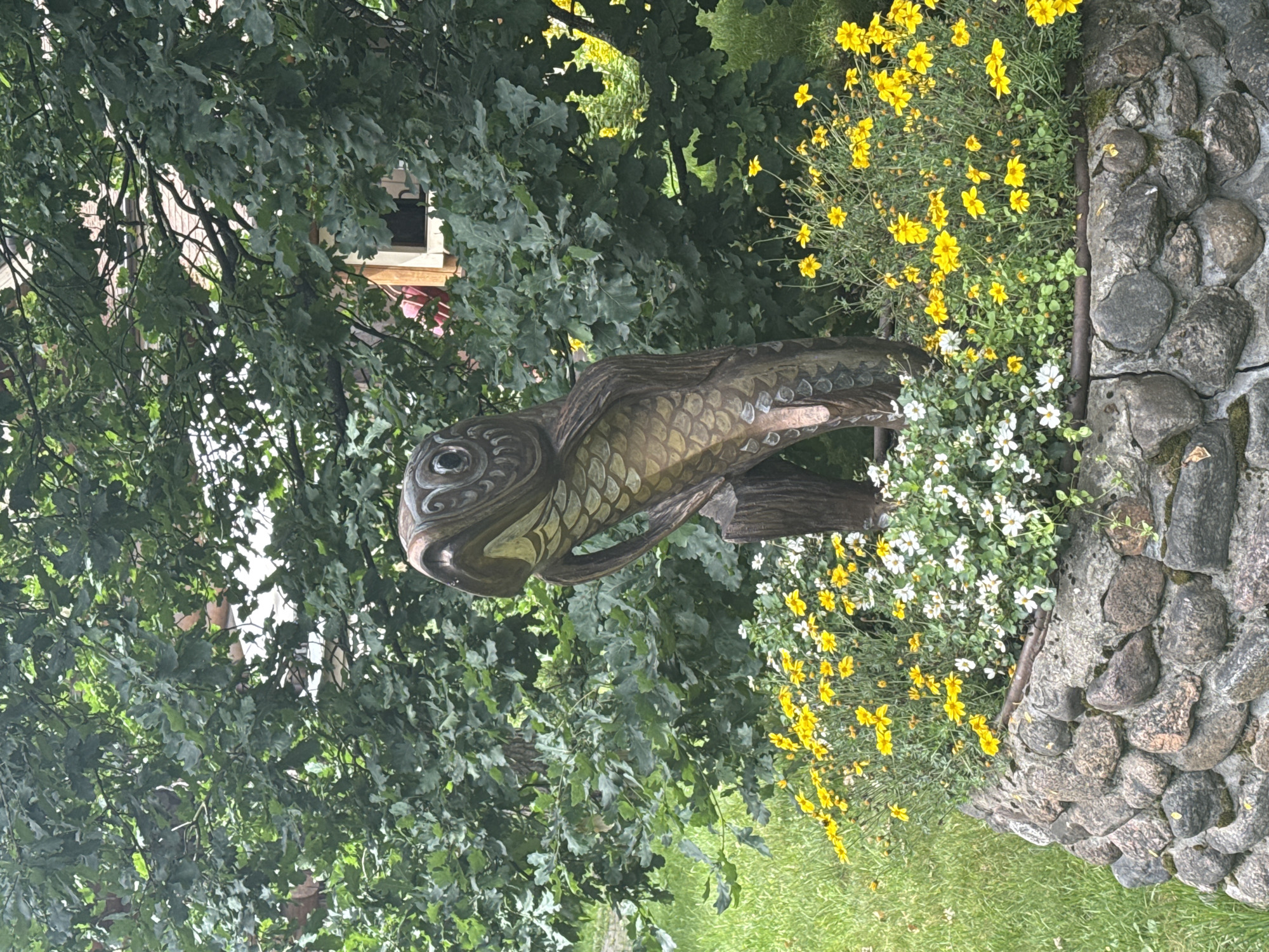 A wooden sculpture of a fish is centrally located in an outdoor setting. The sculpture, which appears to be aged, stands upright and is surrounded by a low, circular stone wall. The wall is filled with a variety of small yellow and white flowers. Behind the sculpture and wall, lush green foliage from trees and bushes dominates the background. Some of the trees have large, lobed leaves. In the upper right portion of the image, glimpses of a building with a red roof and wooden elements are visible through the branches and leaves. The ground is covered in green grass. The scene is set during the daytime, under an overcast sky, as indicated by the diffused lighting and absence of strong shadows. The specific location is identified as Komarovo, Russia, however, no specific landmarks or identifying features of the city are discernible from the image itself. There are no people present in the image.