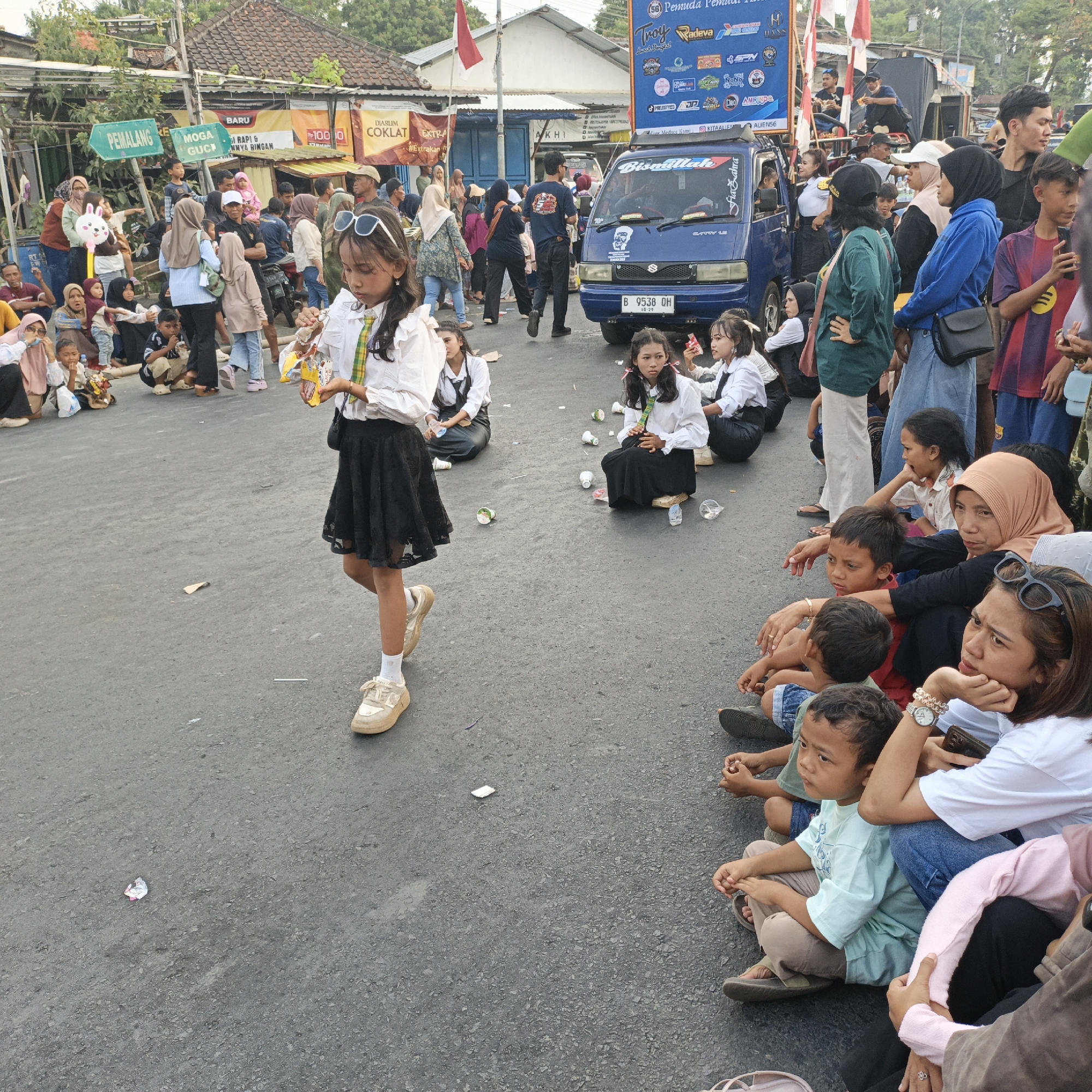 In Randudongkal, Indonesia, an outdoor event is taking place on a paved road. A young girl in a white shirt and black skirt, holding a snack, walks past a crowd of people sitting on the ground. The crowd consists of various individuals, including children and adults, some observing the event and others engaging in conversation. In the background, a blue truck with signage and a red and white flag are visible, suggesting a festive or celebratory atmosphere. The environment appears to be a street, with buildings and overhead wires indicating a populated area. The scene captures a moment of community gathering, possibly a parade or festival, with a mix of participants and spectators.