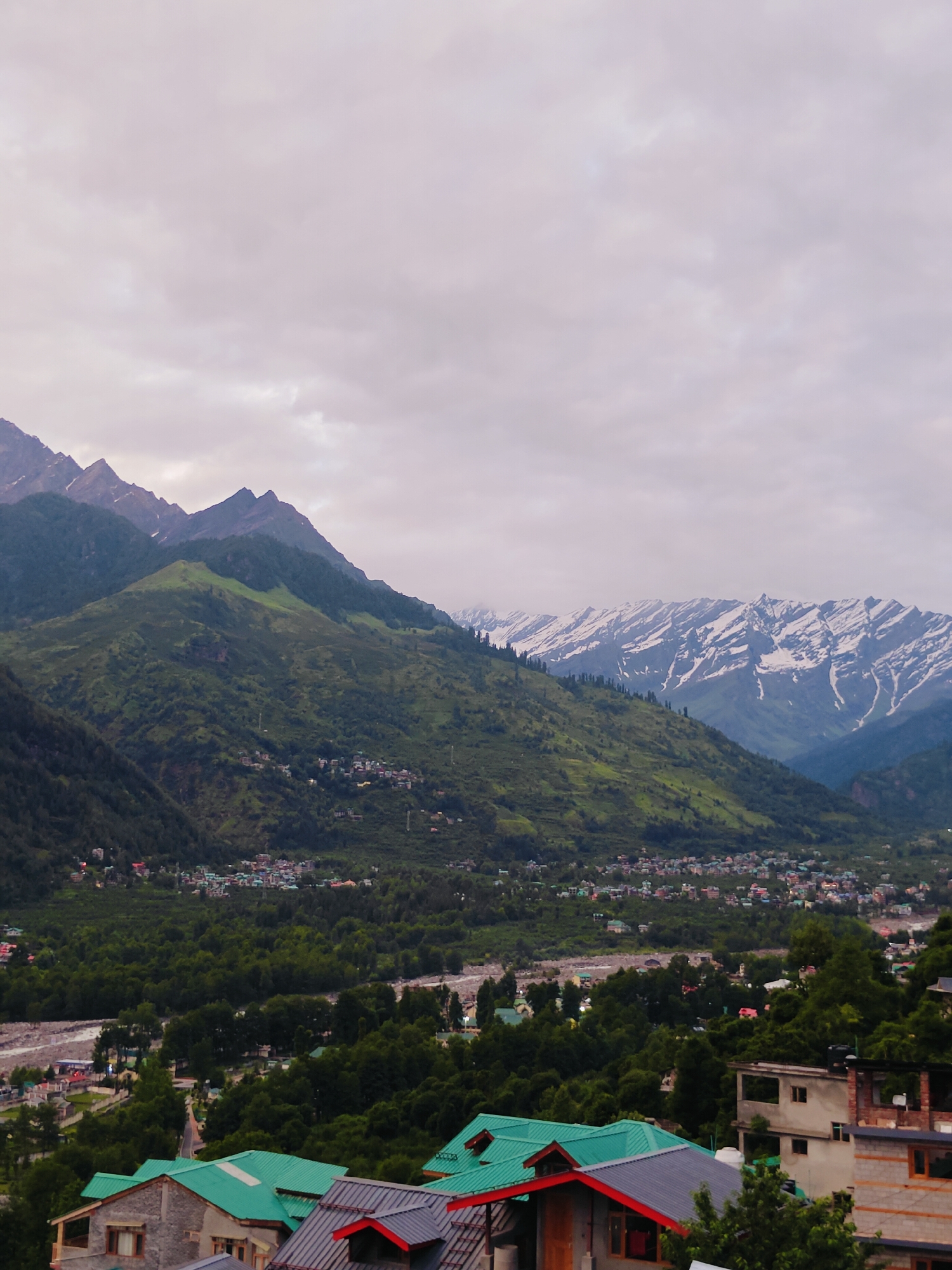 This is a landscape photograph capturing the scenic beauty of Vashisht, India. The image showcases a valley nestled between towering mountains, with a village built on the slopes. The foreground features several buildings with distinct architectural styles, characterized by pitched roofs of varying colors, including green and dark grey. Some buildings are constructed with stone and exposed brickwork. Beyond these structures, the valley unfolds, revealing a dense forest of green trees. A river can be seen winding through the valley. In the mid-ground, the village of Vashisht is visible, with numerous houses and buildings clustered together on the hillside. The backdrop is dominated by majestic mountains, some of which are snow-capped, indicating a high altitude. The sky is overcast with clouds, lending a soft, diffused light to the scene. The overall impression is one of a serene and picturesque mountainous village.