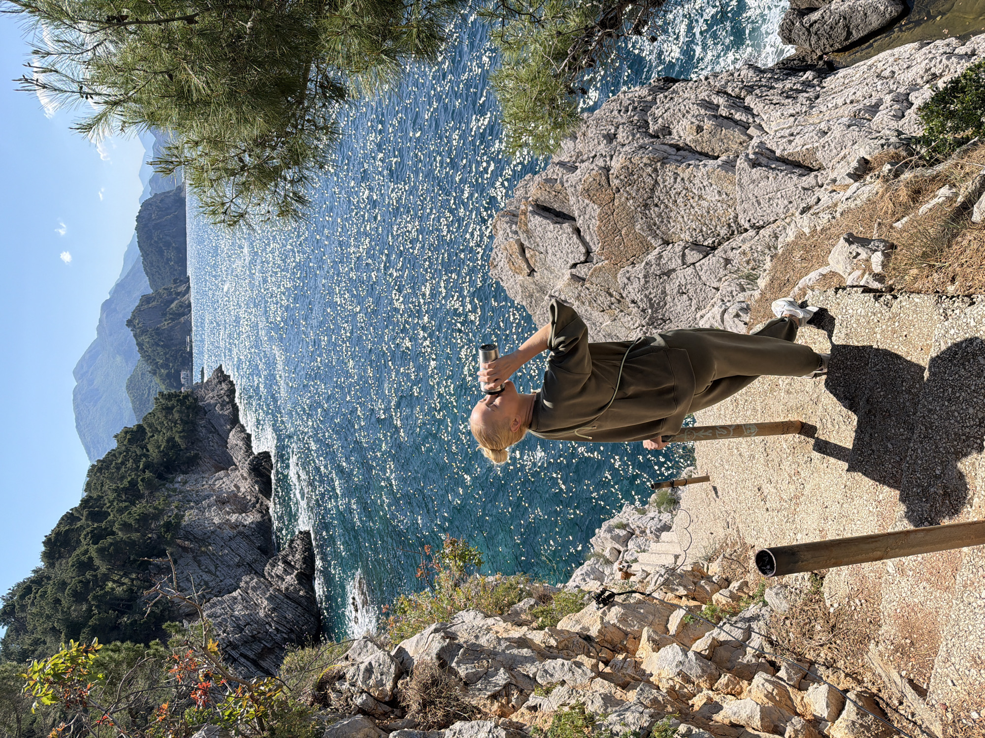 A woman in an olive-green jumpsuit drinks from a silver container on a sunny day in Mirište, Montenegro. The location appears to be a rocky cliff overlooking the Adriatic Sea. The water sparkles in the sunlight and mountains are visible in the distance. The foreground shows vegetation and a rough path with metal railing.
