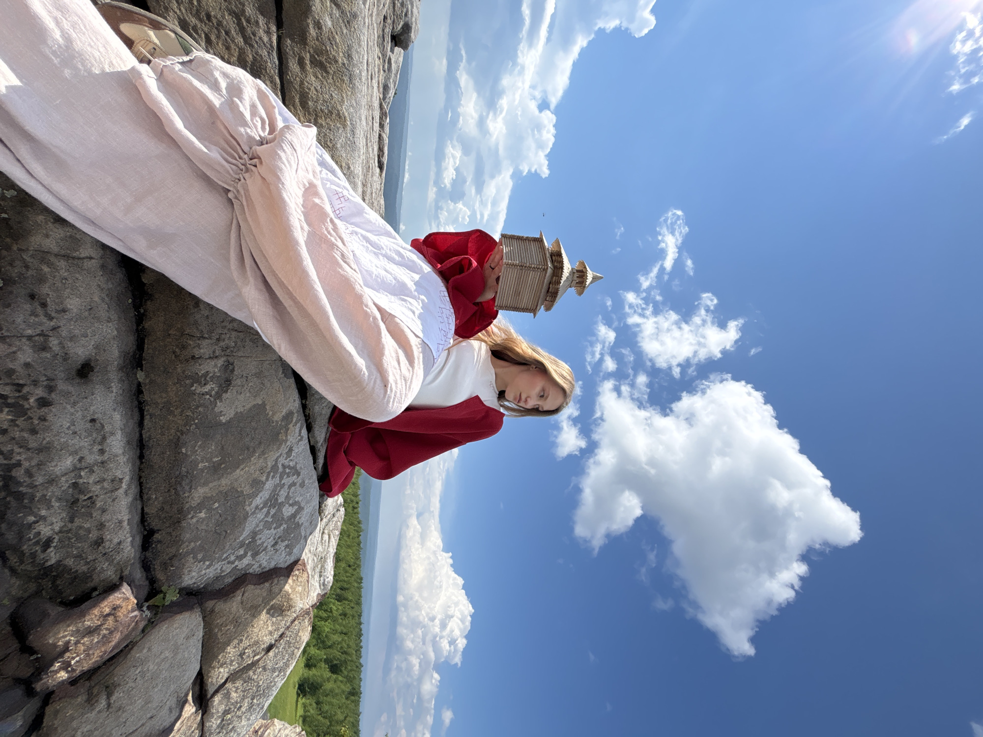 A young woman with long blonde hair sits on large rocks on a sunny day. She is wearing a white top, a red jacket, and a long, flowing pink skirt. In her hands, she holds a small wooden model of a building. The background shows a bright blue sky with fluffy white clouds and a distant green forest. The location is the Kuberla rock formation, also known as the "Devil's Finger," in Gubakha, Russia, a popular tourist attraction and natural landmark.