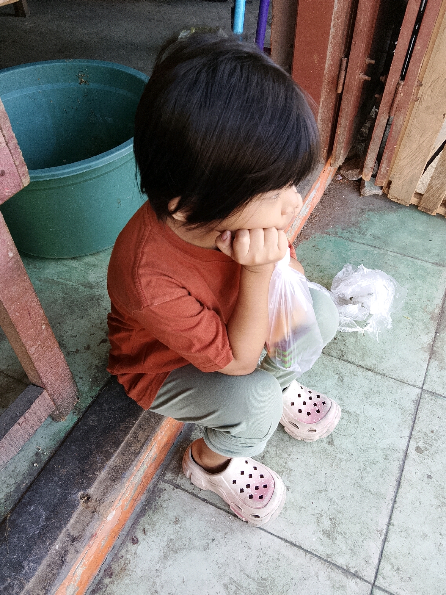 A child with dark hair sits on a tiled floor, facing to the right. The child wears an orange t-shirt and light green pants, with light pink clogs on their feet. Their right hand rests on their chin, and they hold a plastic bag containing items. A second crumpled plastic bag is nearby. To the left, a large green bucket is partially visible. The background features wooden structures and a rusty metal door. The scene appears to be outdoors, possibly in a yard or alleyway, consistent with a location in Talise, Indonesia.
