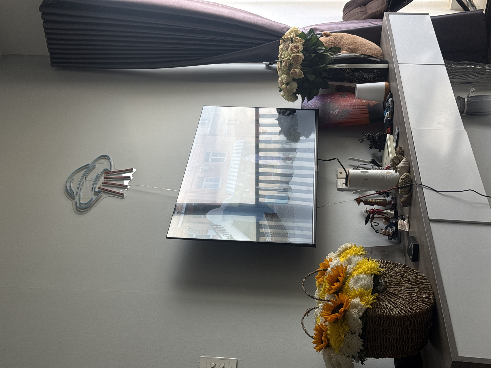 The image depicts an indoor scene, likely a living room, set in Nha Trang, Vietnam. A large, flat-screen television is mounted on a grey wall. Below the television is a console table with various decorative items, including a large woven basket filled with yellow and white flowers, a vase with a bouquet of pale pink roses, and several small figurines. The overall impression is one of a tidy and decorated living space.