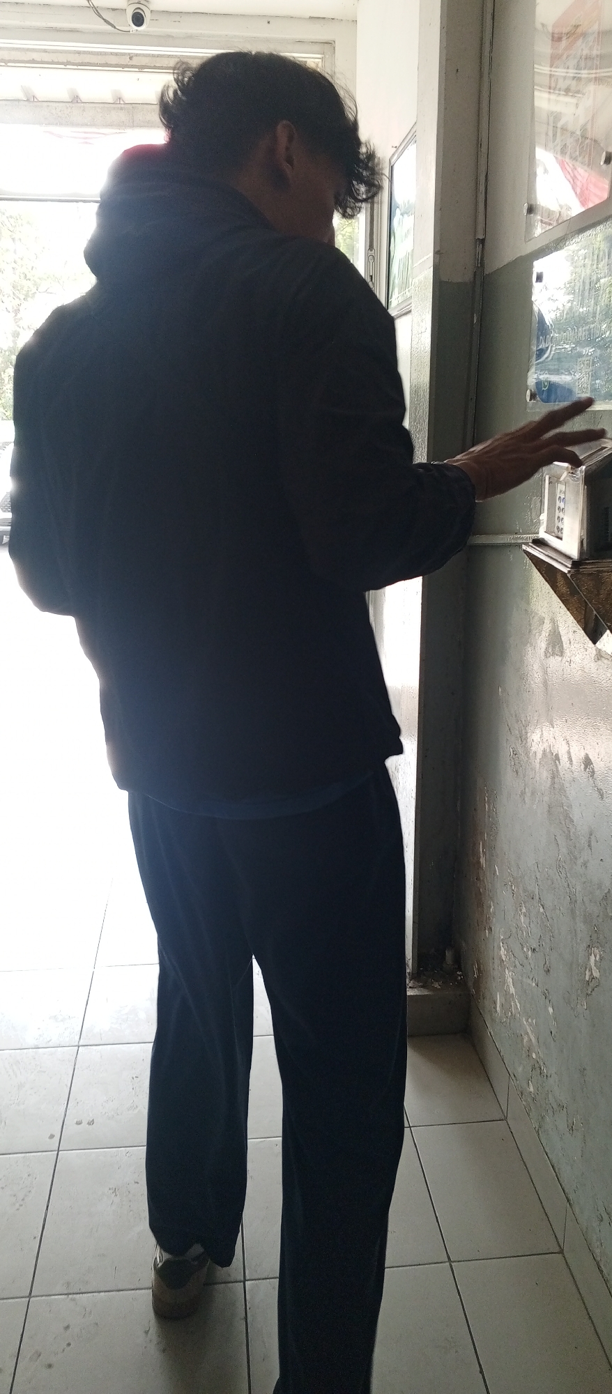 A person wearing a black jacket and dark pants is standing and reaching towards a clear plastic device with buttons. The device is mounted on a wall. The person's left arm is extended, and their fingers are visible near the buttons of the device. The background shows a tiled floor and a weathered wall with peeling paint. There is a CCTV camera visible in the upper left corner of the image. The scene appears to be indoors, possibly in a public or commercial area.
