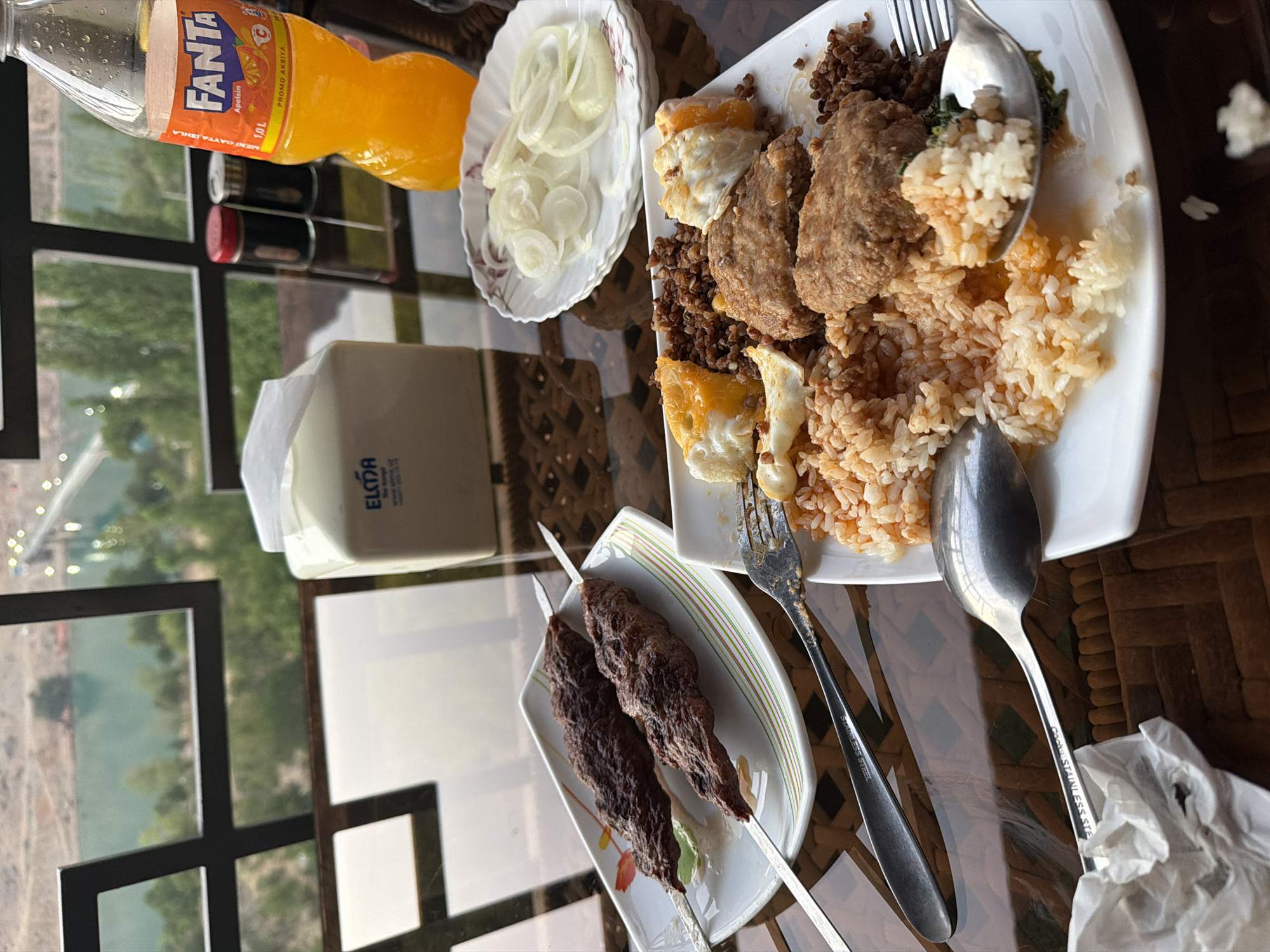 The image depicts a table setting at an outdoor restaurant overlooking a body of water and a bridge in the distance. The table is laden with a meal, including two skewers of grilled meat, a plate of rice with meat patties and fried eggs, and a side of sliced onions. A bottle of Fanta soda is also visible, along with a box of tissues and salt and pepper shakers. The overall scene suggests a casual dining experience. The view of the water and bridge suggests a location with a scenic backdrop, possibly a lakeside or riverside restaurant. The presence of local elements like the food preparation and the overall ambiance could indicate a restaurant in Nanay, Uzbekistan, known for its natural beauty and hospitality. The lighting suggests it is daytime, possibly late afternoon or early evening, with the sun casting a warm glow on the scene.