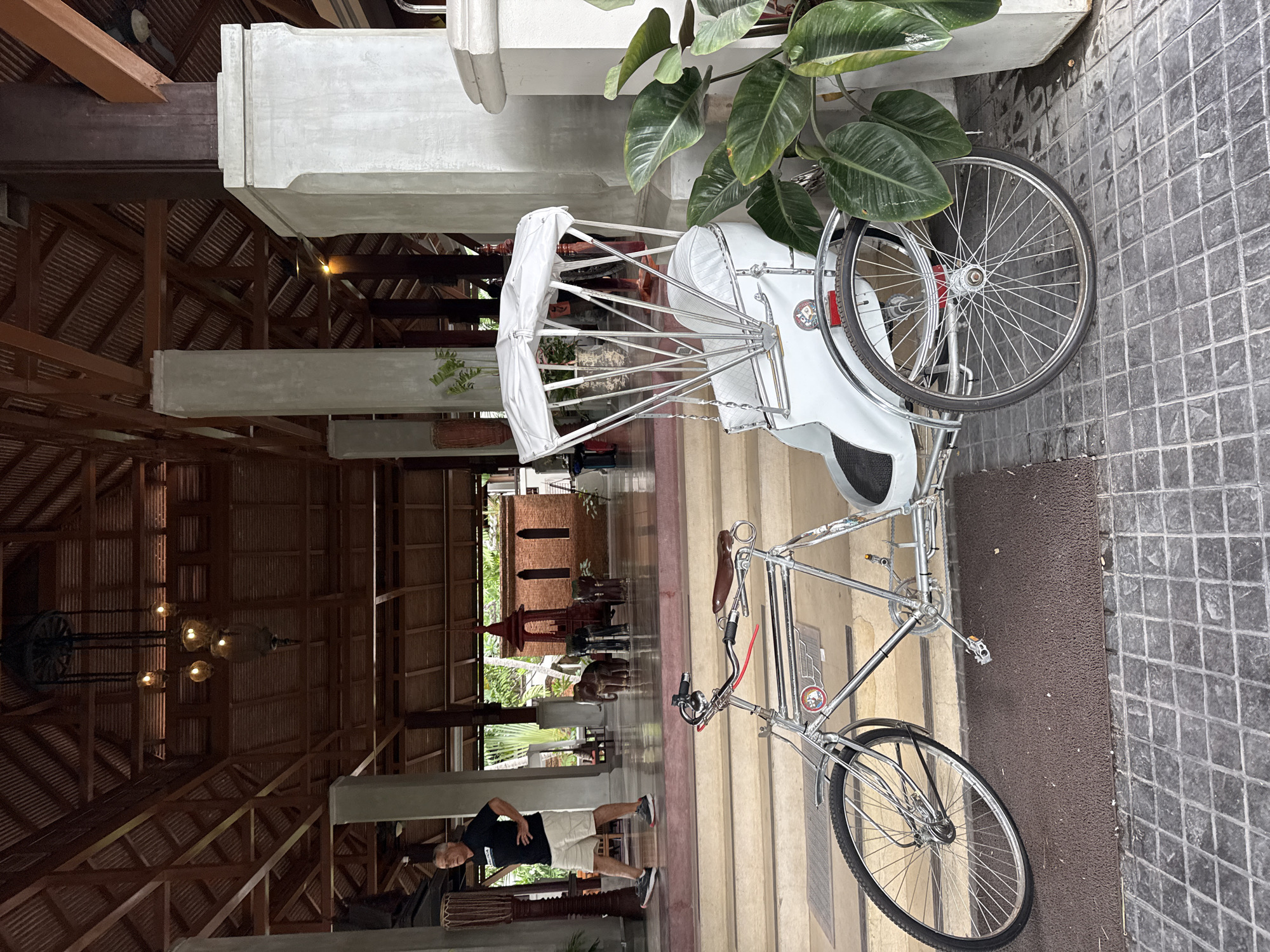 The image depicts a tricycle with a white canopy parked on a tiled floor in front of a set of stairs. The tricycle is a classic model with a chrome frame and leather-look seat. Behind the tricycle, a man in a black t-shirt and shorts walks up the stairs. The background shows the interior of a building with a wooden roof structure, decorative pillars, and some plants. Further in the background, there are elephant statues and an ornate brick structure. The scene appears to be an outdoor area of a hotel or resort in Ko Samui District, Thailand, given the architectural style and the presence of tropical plants. The overall atmosphere is one of a warm, sunny day.