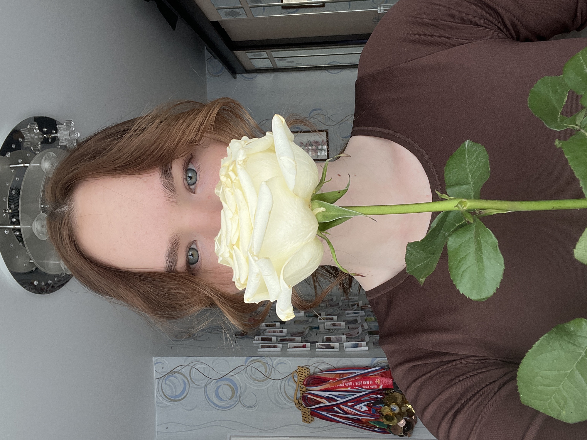 A young woman with light brown hair and blue eyes is holding a white rose in front of her face. She is wearing a brown long-sleeved shirt. In the background, a shelf displays small items and medals with ribbons. A silver ceiling light fixture is visible on the left. The location appears to be indoors, possibly in a home in the city of Chaikovsky, Russia, based on the inscription visible on one of the medals.