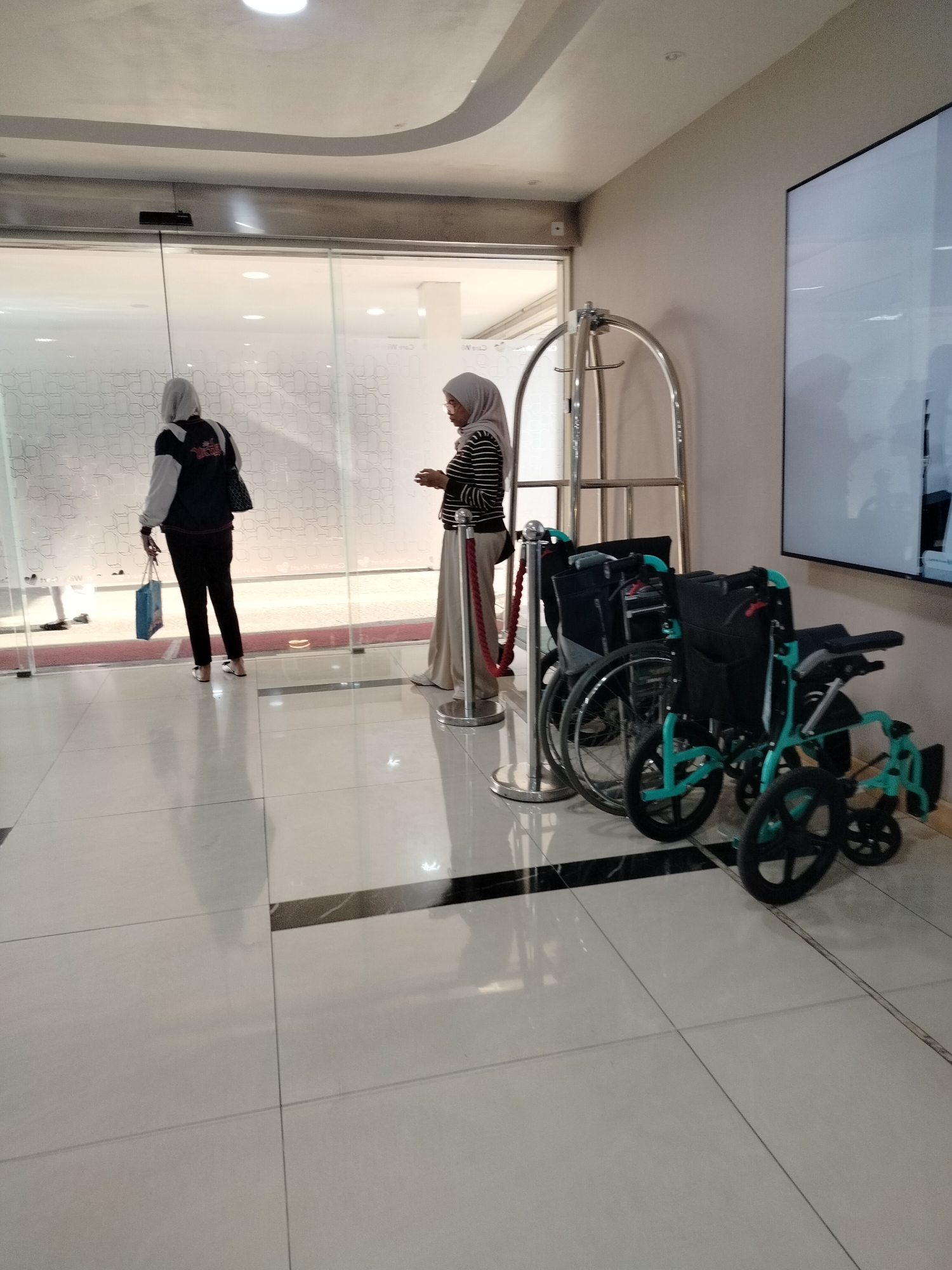 Two women are standing inside a building in East Jakarta, Indonesia. One woman, wearing a hijab, dark pants, and a jacket with white sleeves, is holding a blue shopping bag and standing near a glass door. The second woman, also wearing a hijab and a striped shirt with a long skirt, is standing further inside the building, next to a red rope stanchion. In the foreground, two wheelchairs are parked. The floor is tiled with a reflective surface, and the walls are a neutral color. A luggage cart is visible behind the women. The lighting suggests an indoor environment, possibly a hotel or a commercial establishment.