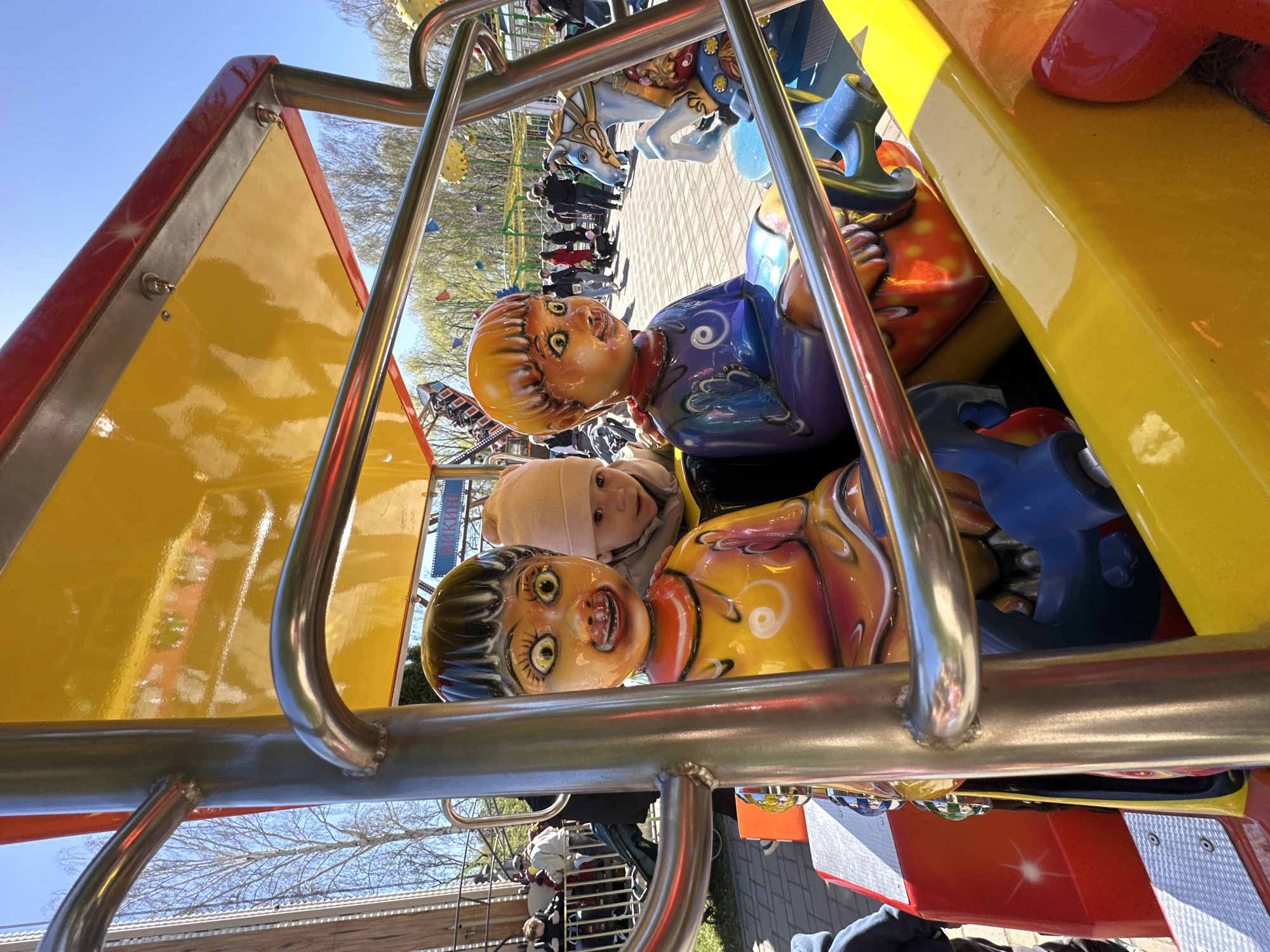 The image captures a scene from an amusement park ride. A young child, wearing a beige beanie, is seated on a carousel-like attraction with brightly colored, cartoonish figures, including two distinctive dolls with exaggerated facial features. The ride has a yellow and red canopy and is constructed with stainless steel bars. In the background, a crowd of people can be seen lining up, along with a glimpse of a roller coaster. The location is identified as the city of Yekaterinburg, Russia, evidenced by signage in Russian.