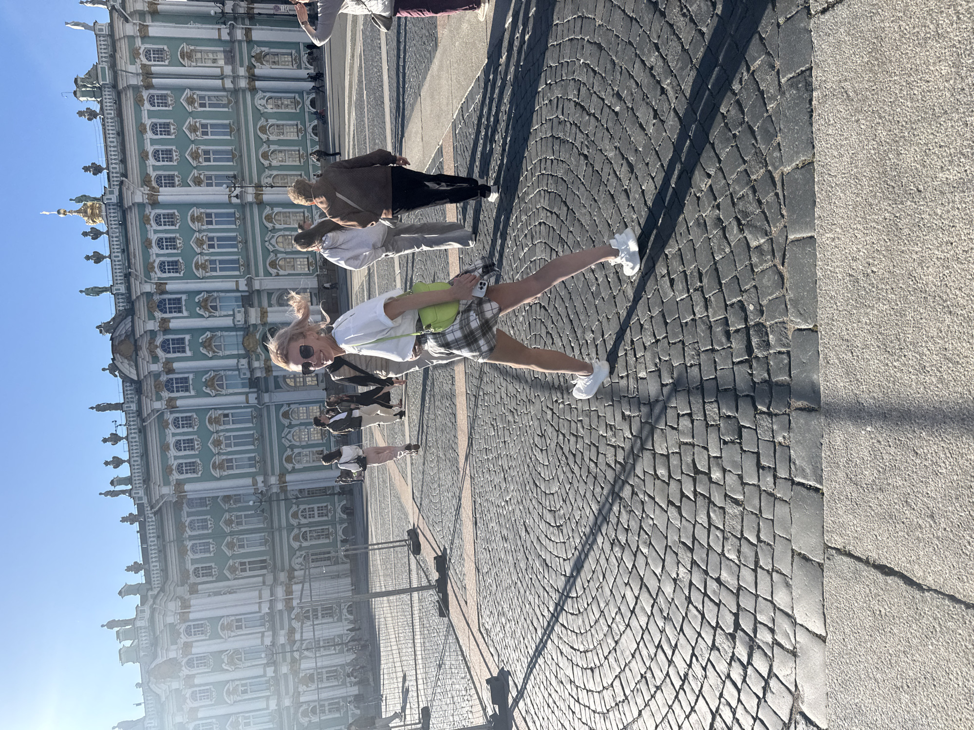 A young woman with blonde hair and sunglasses is walking on a cobblestone square in front of the State Hermitage Museum in Saint Petersburg, Russia. She is wearing a white t-shirt, a plaid skirt, and white sneakers, and carrying a bright green bag over her shoulder. She is looking towards the camera and smiling. Several other people are walking in the background, as well as scaffolding and fencing in front of the museum. The sky is clear and blue, indicating a sunny day.