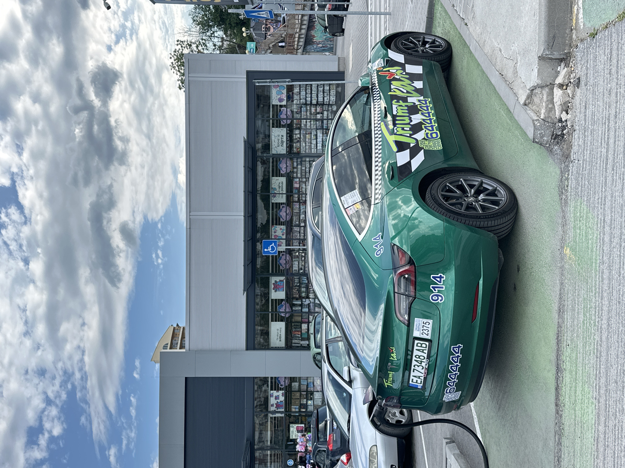 The image captures a green Tesla Model 3 parked in front of a store. The car is customized with the logo of "Triumf Taxi" and the phone number "644444" prominently displayed. The license plate reads "EA 7348 AB," and the number "914" is marked on the car's rear. The car is in a parking space designated for electric vehicles, charging with a cable plugged into it.
The store behind the car appears to be a general goods store, with items visible through the glass front. A sign indicating accessibility for people with disabilities is displayed on the storefront.
The sky above is partly cloudy. In the background, there are signs of other buildings and infrastructure. The location is in Veliko Tarnovo, Bulgaria, as indicated by the license plate information.