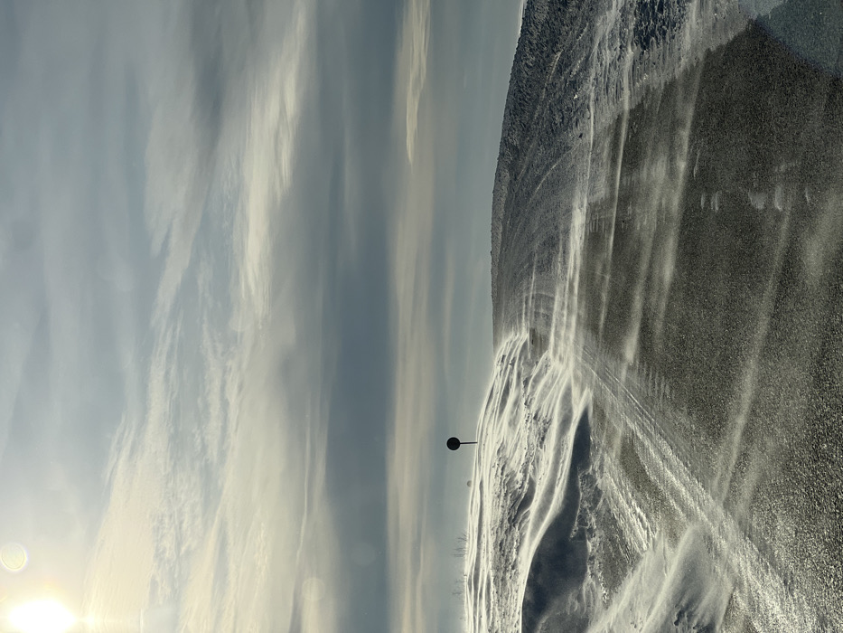 The image depicts a winter road scene in Ol'khovo, Russia, under a partly overcast sky with the sun visible in the upper-left quadrant, producing a lens flare. A dark, presumably asphalt, road surface extends from the foreground into the mid-ground, exhibiting tire tracks. The road and surrounding terrain are partially obscured by actively blowing snow, indicative of strong winds, which sculpt the snowdrifts to the left. A dark, circular sign mounted on a pole stands on the snowdrift to the left of the road, with minimal bare vegetation visible behind it. To the right, a snow-covered embankment rises, showing interspersed darker patches of terrain. No individuals are visible within the frame.