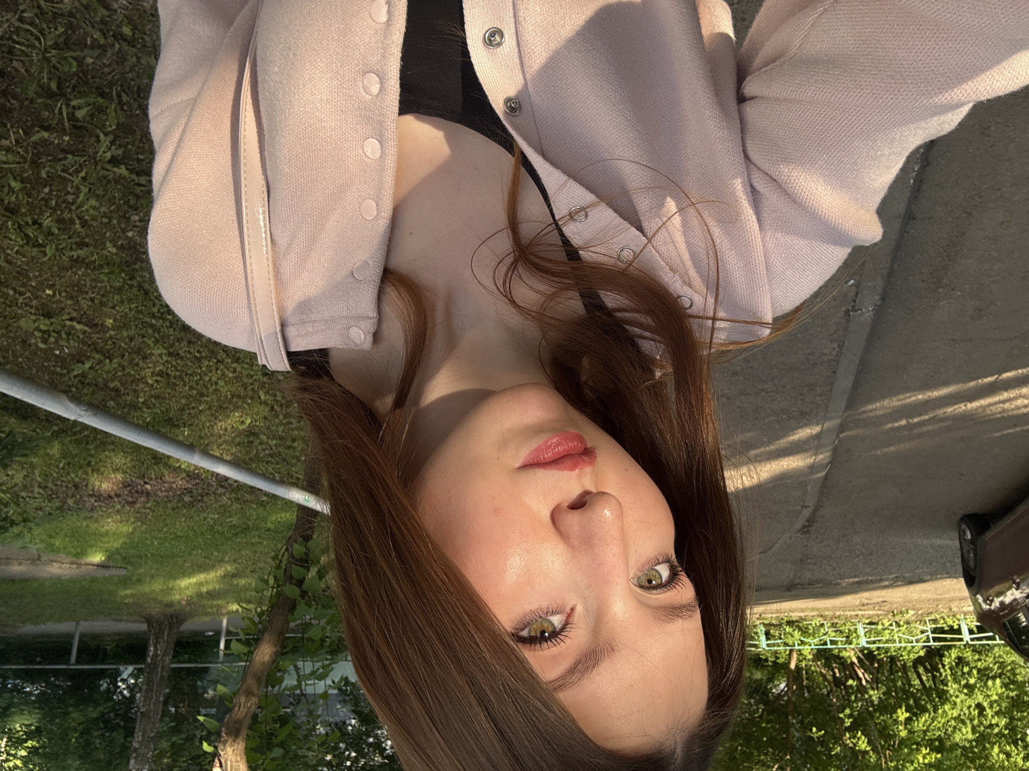 This image captures a close-up selfie of a young adult female located in the city of Chaikovsky, Russia, during what appears to be late afternoon or early evening, characterized by warm, directional sunlight and long shadows.
The subject, a female with long, straight to slightly wavy brown hair parted centrally, occupies the majority of the frame. She has hazel-green eyes with prominent eyelashes, a small nose piercing on her left nostril, and a small, reddish mark or blemish on her right cheek. Her lips are lightly colored with a reddish-pink hue. She is wearing a light pink, possibly knit, cardigan fastened with multiple silver snap buttons down the front. Beneath the cardigan, a black top or camisole is visible, featuring a V-neckline. The strap of a light-colored (possibly cream or pale pink) shoulder bag is visible on her right shoulder. She is looking directly towards the camera with a neutral to slight smile expression.
The background consists of an outdoor urban-park environment. To the left, an asphalt or paved path, light gray with visible cracks and texture, extends into the distance. Long, diagonal shadows are cast across this path. A dark-colored (possibly brown or maroon) car, either a sedan or small SUV, is partially visible parked on the left side of the path. Behind the path and car, dense green foliage, including trees and bushes, is present, interspersed with a light blue or greenish metal fence featuring decorative curves. To the right of the subject, a grassy area with varied shades of green and some exposed dirt is visible, along with the lower trunks of several trees, some with light bark. A silver-colored metal railing or fence section runs horizontally across the mid-ground on the right side of the image, disappearing behind the subject. The overall lighting is warm, with direct sunlight highlighting the subject's face and hair, creating distinct shadows.