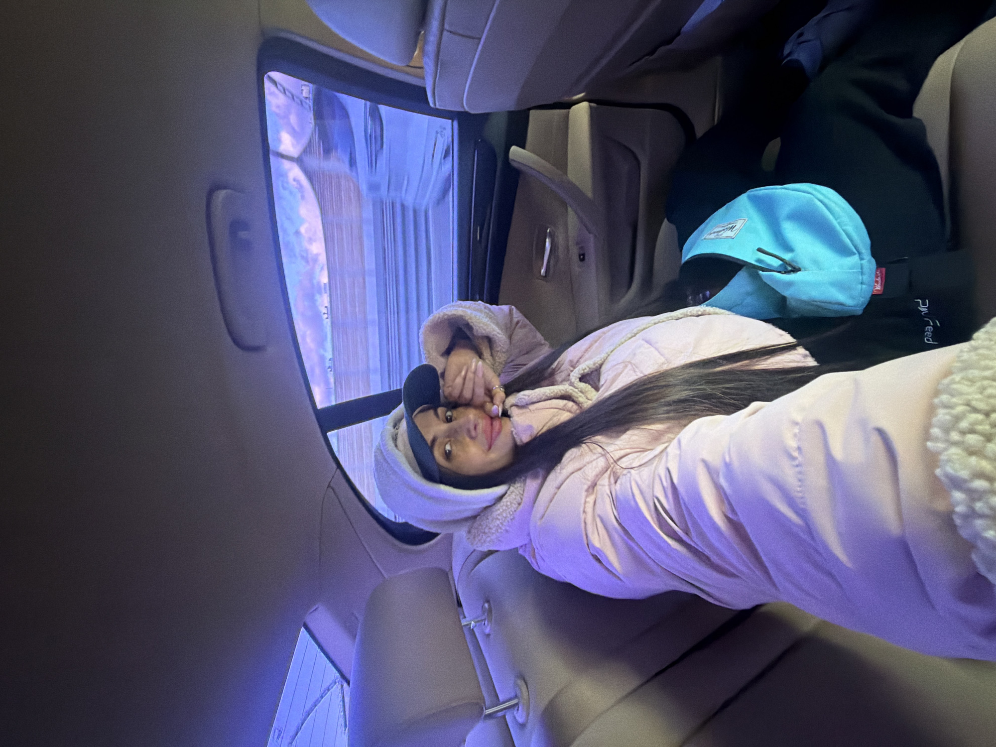 A young woman is lying down in the back seat of a car. She has long, dark hair and is wearing a light pink hooded jacket. She is looking at the camera with her right hand near her face. To her right is a blue Herschel brand backpack. The car interior is beige. Out the car window, a street scene with vertical lines is visible, potentially suggesting Saint Petersburg, Russia based on architectural styles.