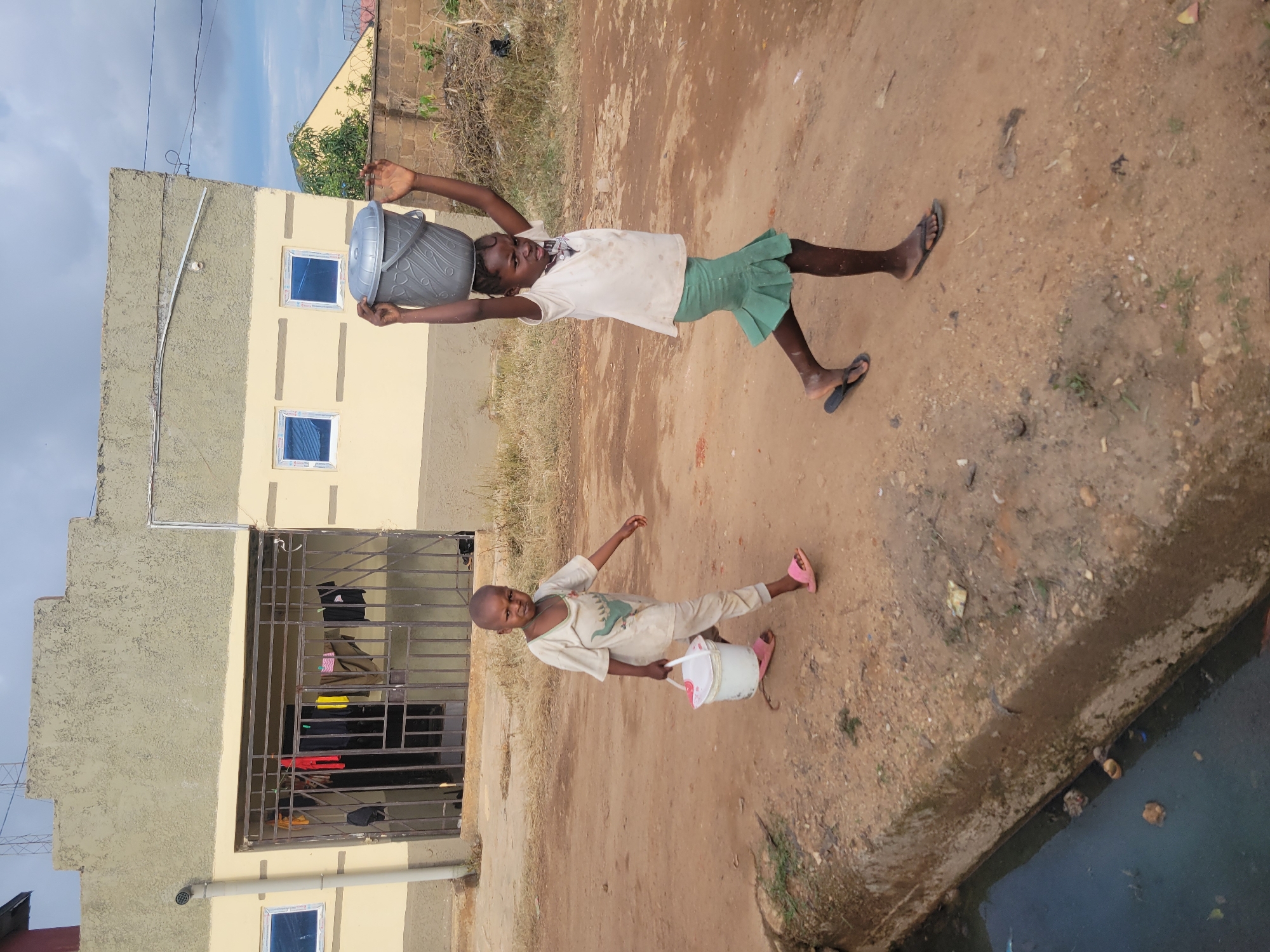 The image depicts two children on a reddish-brown dirt road in the city of Jos, Nigeria, against a backdrop of local architecture under a partially overcast sky.
In the mid-ground, a young girl, estimated to be between 8 and 12 years old, walks from left to right. She is carrying a large, lidded grey plastic bucket balanced on her head, steadying it with both hands raised. She wears a light-colored t-shirt, a green pleated skirt, and dark flip-flops. Her dark skin is visible on her arms and legs, and her expression appears to show effort.
Further left and slightly closer to the foreground, another child, appearing younger (approximately 5-8 years old), walks from right to left. This child carries a smaller white plastic bucket with a pink handle in their left hand. They are dressed in a light-colored, possibly stained, long-sleeved top featuring a green dinosaur graphic, and light-colored trousers, along with pink flip-flops. Their facial expression is a slight frown.
The immediate foreground on the left features a concrete-lined open drainage ditch containing murky, dark water with visible debris and trash. The dirt road itself is uneven and dusty.
In the background, a single-story building with a light-yellow painted facade and a textured grey upper section dominates the left side of the frame. A dark metal security gate covers the main entrance, with several pieces of multi-colored laundry (including red, yellow, pink, and black items) hanging to dry on its bars. Two small windows with blue-tinted panes and white frames are visible on the building's facade to the left of the gate, and two similar windows are seen on the right side of the building. A light-colored vertical pipe runs down the left exterior corner. Utility lines are visible against the sky above the building. Further to the right, beyond some dry brush, another building or structure with a lighter-colored roof is partially visible. The sky is largely covered by grey clouds with some patches of lighter blue visible.