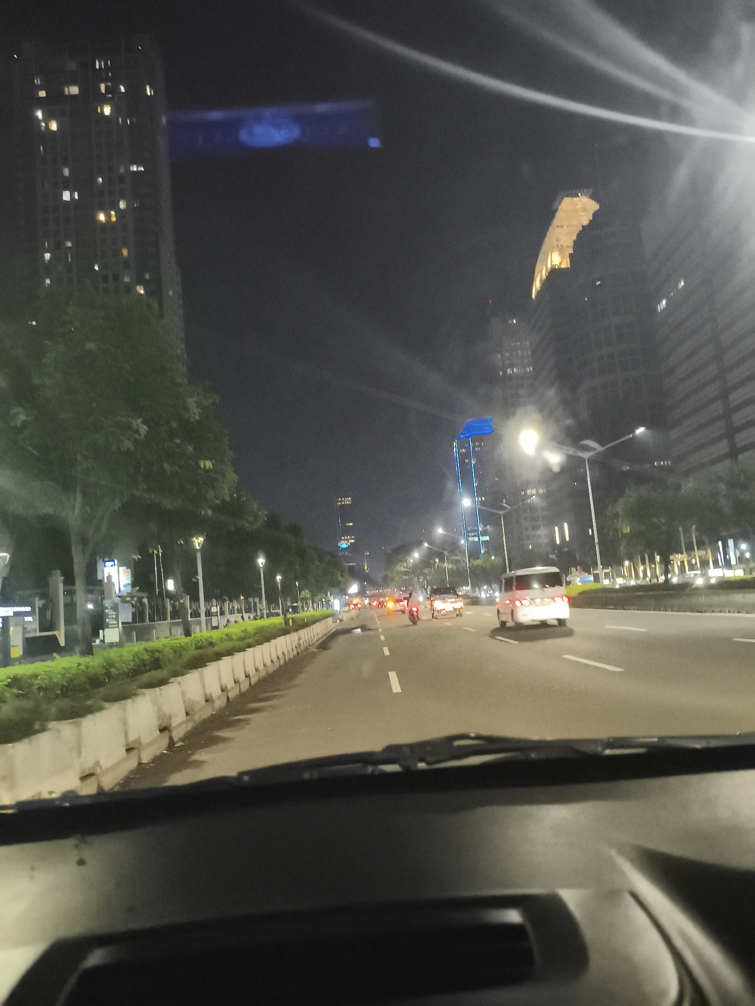 The image depicts a night scene from inside a vehicle traveling on a city road in Unknown, Unknown. The road has multiple lanes and is lined with concrete barriers on the left side, followed by a row of greenery. Streetlights illuminate the road and surroundings. A variety of vehicles, including cars and motorcycles, are visible on the road ahead. In the background, tall buildings rise into the night sky, some of which are lit up. The view is partially obstructed by the car's dashboard and windshield.
