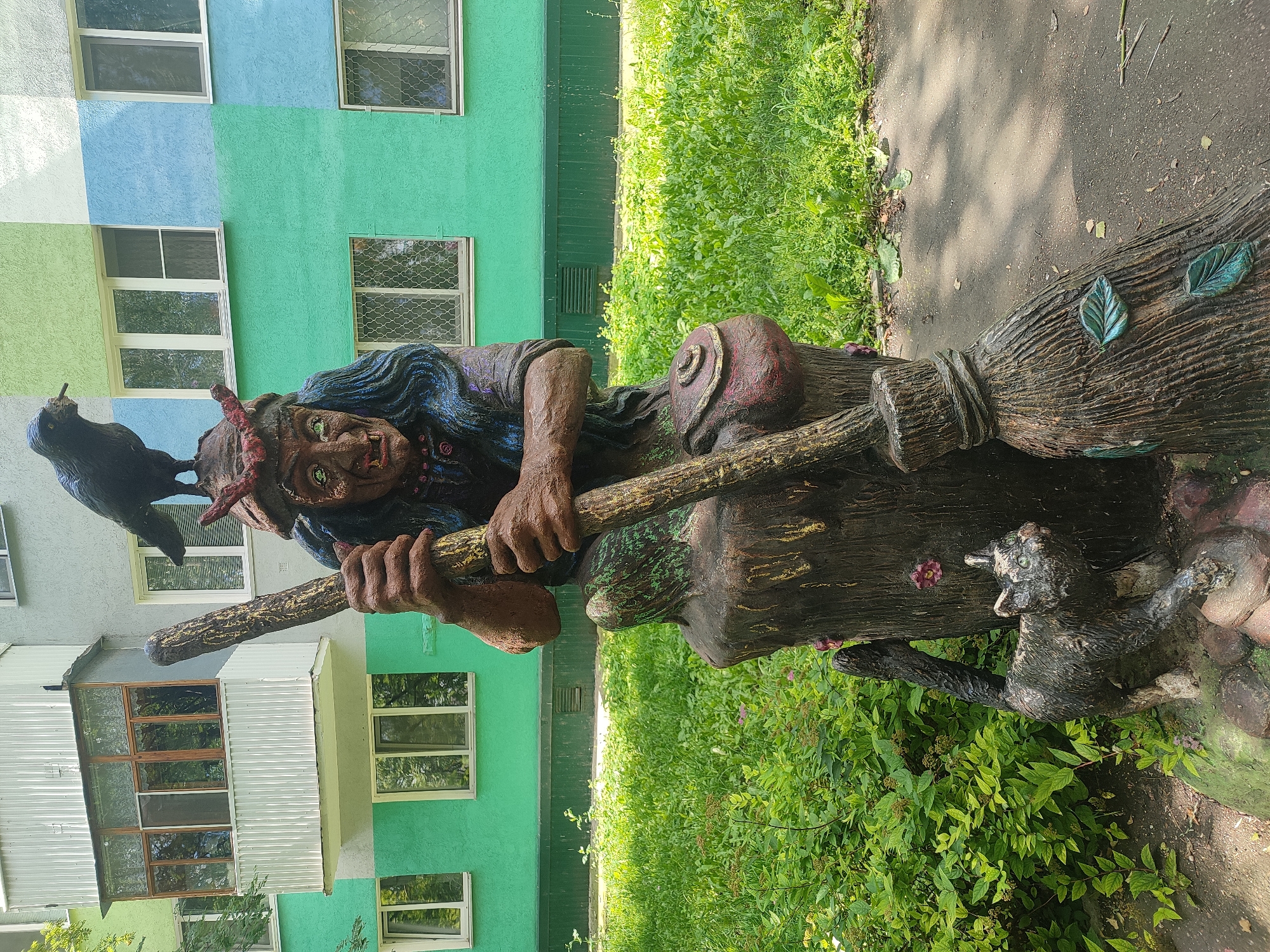 The image shows a statue of a witch sitting on a stump, holding a broomstick. A black bird is perched on her head, and a black cat is at her feet. The statue is located in front of a colorful apartment building in Naberezhnye Chelny, Russia. The building has a balcony on the second floor and windows on the upper floors. The ground in front of the building is covered with grass and some scattered leaves. The statue is made of a material that appears to be painted concrete or plaster, and it is detailed with a witch's face, hair, and clothing, as well as the bird and cat. The broomstick is also detailed with what appear to be leaves. The scene is set in an outdoor environment during the daytime.