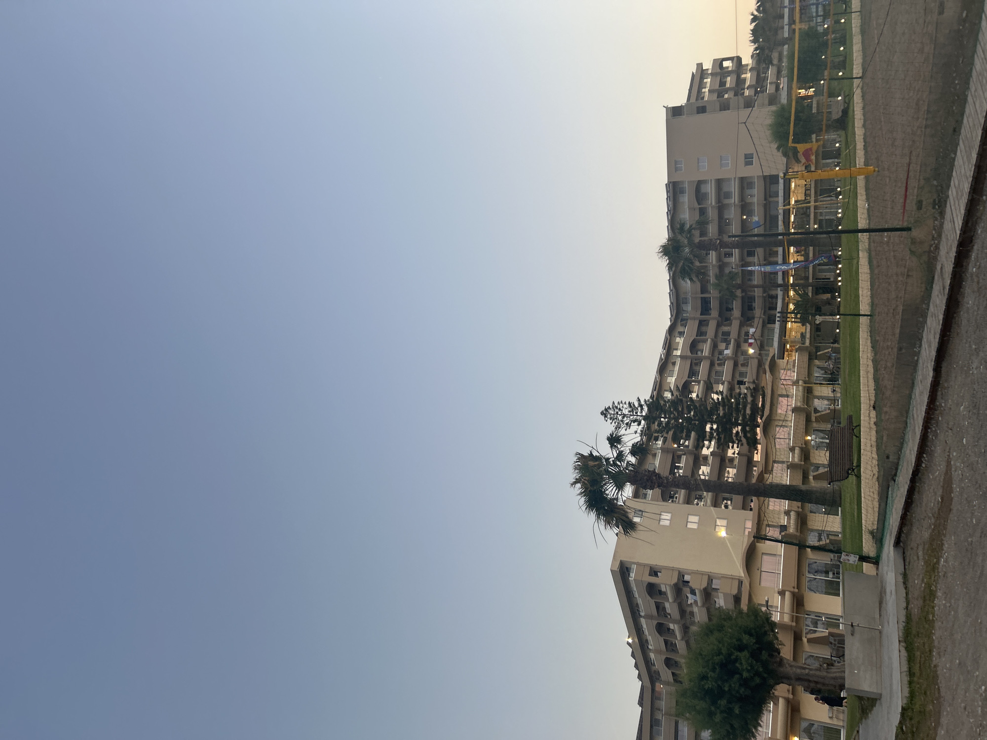 This is a vertically oriented, outdoor, eye-level photograph of a resort complex, likely in Rhodes, Greece. The sky takes up the majority of the left side of the frame and is a pale blue, transitioning to a light yellow near the horizon on the right, indicating it is either dawn or dusk.
The primary subjects are several multi-story resort buildings with light-colored facades. These buildings appear to be modern hotels or apartment complexes, with numerous balconies and windows visible. A few palm trees and other lush greenery are interspersed around the buildings and in manicured lawns.
In the foreground on the right side of the image, a sandy area is visible, suggesting a sports court, possibly for beach volleyball, as indicated by a net and yellow poles. A paved walkway runs alongside this area.
There are no clearly discernible people engaged in any specific activity, though a few figures might be present in the lower parts of the buildings, too indistinct to be sure. The overall mood is peaceful and serene, enhanced by the soft, natural light of the transitional time of day.
No visible text is present in the image. The scene depicts a tranquil resort setting in a coastal or near-coastal location, likely a tourist destination given the architecture and amenities.