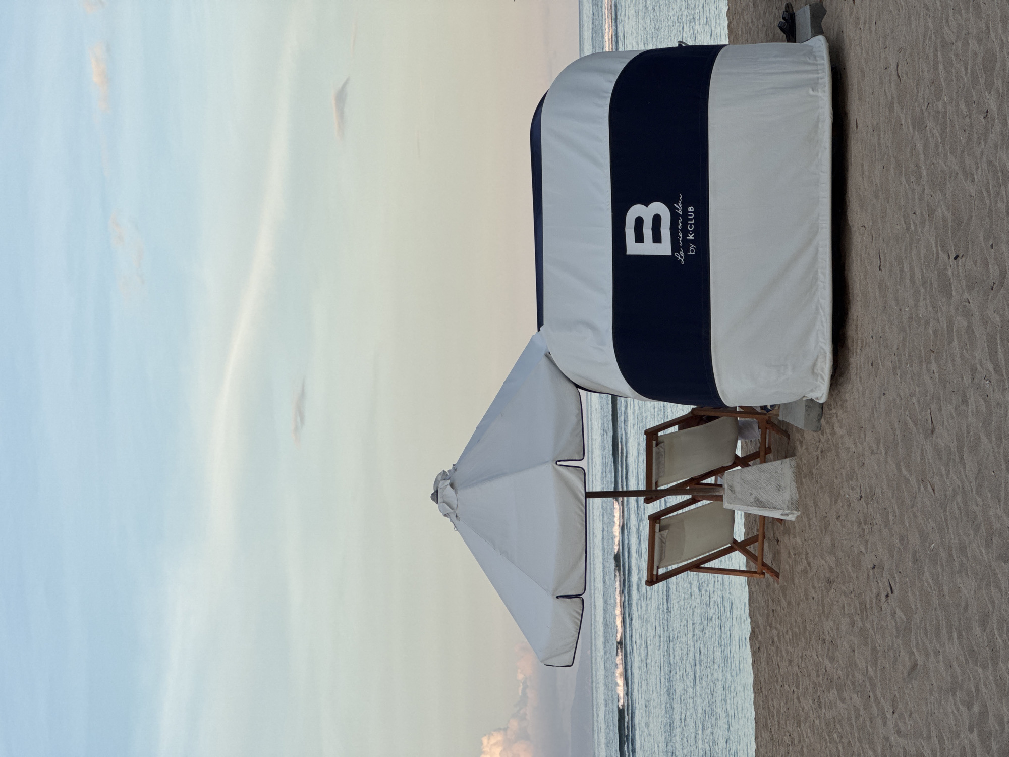 The image depicts a beach scene. There's a covered lounge area with a white and navy blue striped design, featuring the letter "B" and the words "La vie en bleu by K-CLUB". Two wooden beach chairs are positioned in front of the lounge. An umbrella provides shade. In the background, the ocean meets a sky with soft colors, possibly during sunrise or sunset. The location is Unknown, Unknown.
